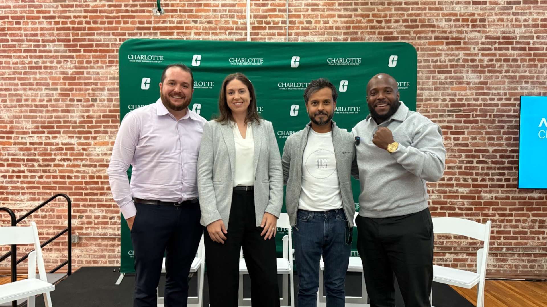 Four people standing side by side smiling in front of a green backdrop with "CHARLOTTE" logos, against a brick wall. Two men wear light gray jackets; woman in gray blazer; one man in light purple shirt.