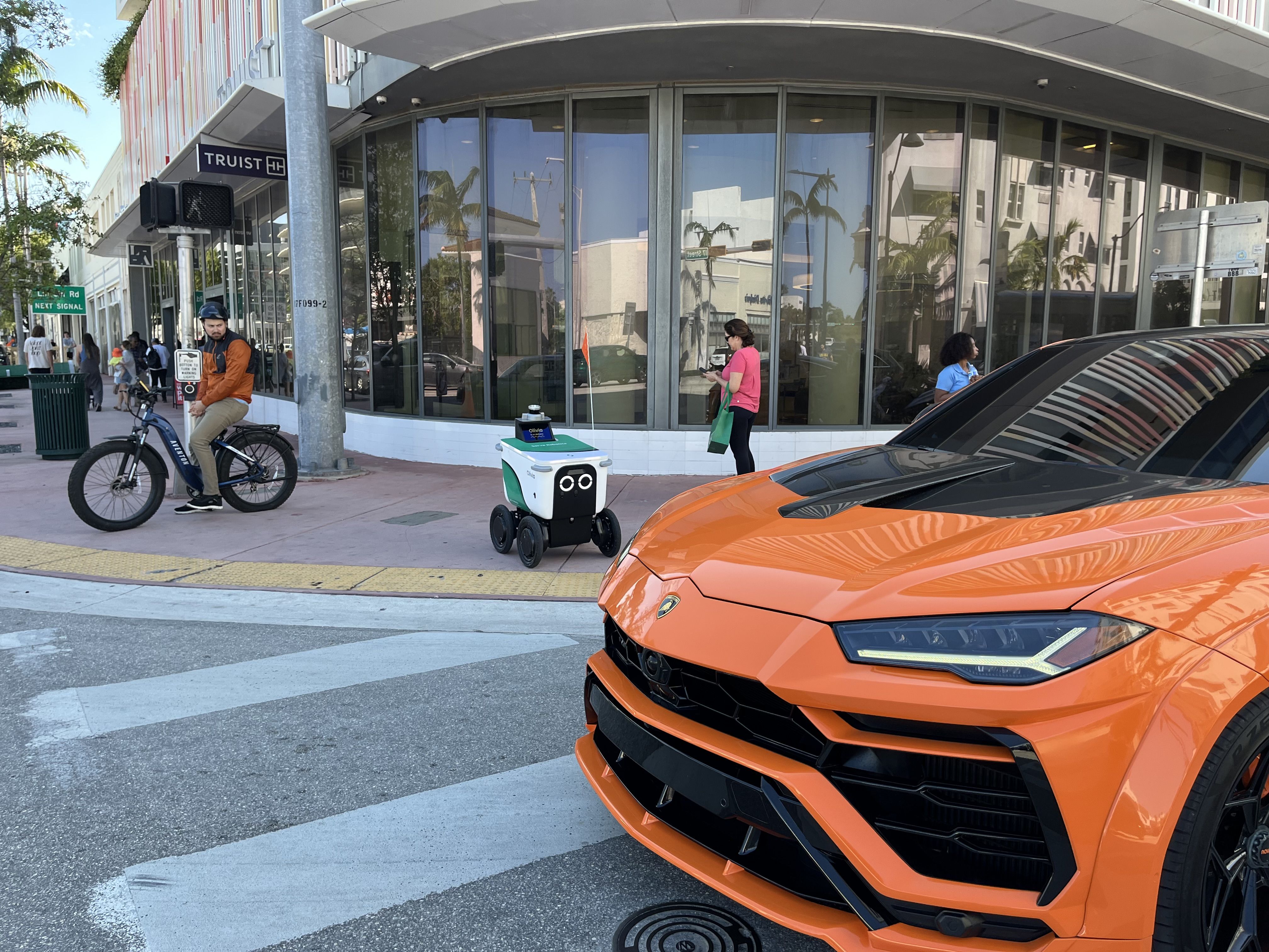 A Lamborghini blocks the crosswalk as a Serve Robotics delivery robot waits to cross the street.