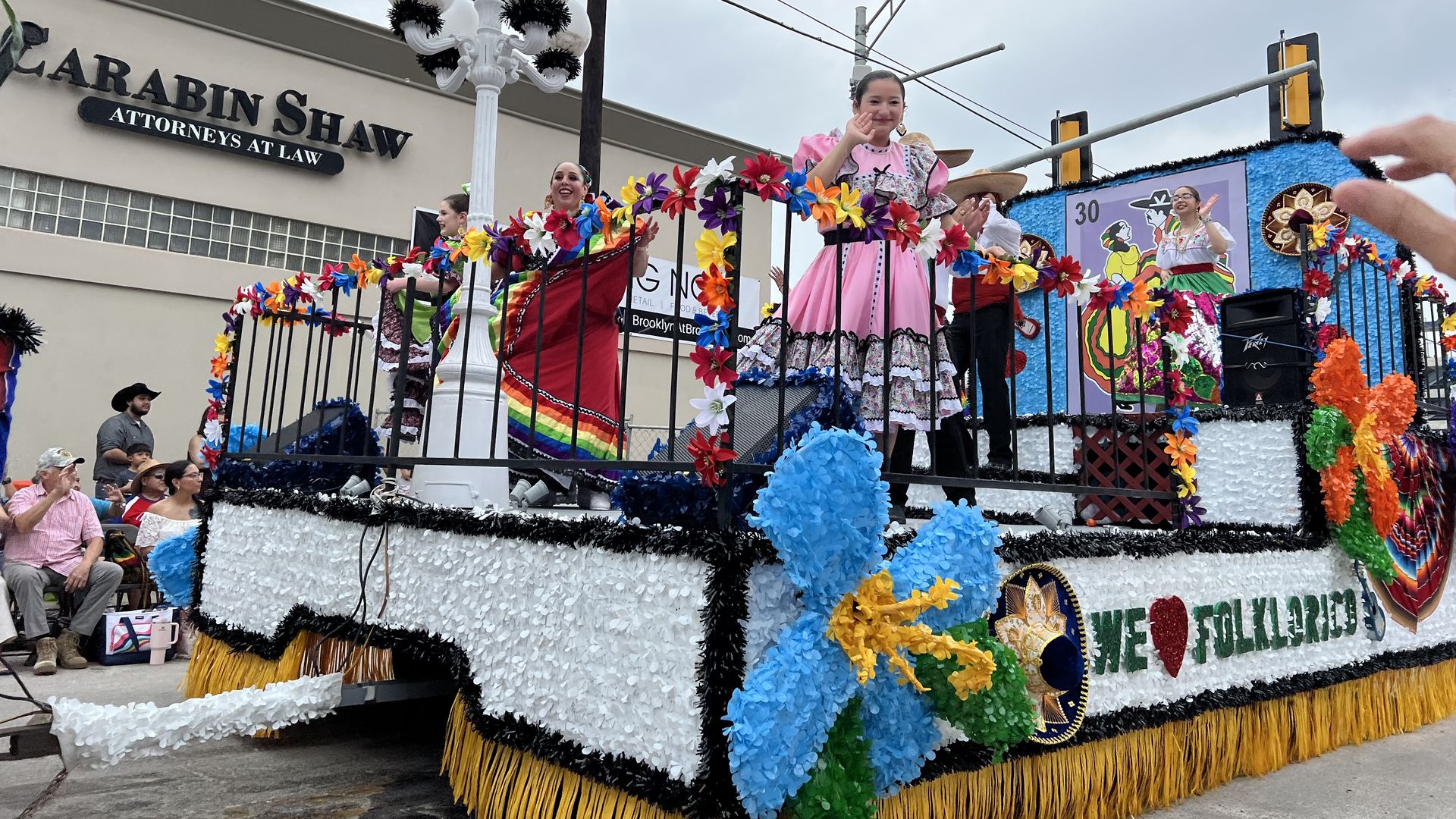 Colorful parade float with dancers in bright dresses, flower garlands, and fringe; spectators watch as a building sign reads "Carabin Shaw Attorneys at Law" in the background.