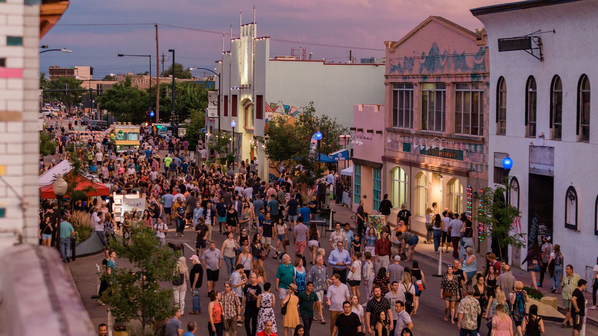Crowded street festival a with people walking along decorated buildings featuring murals and colorful lights.