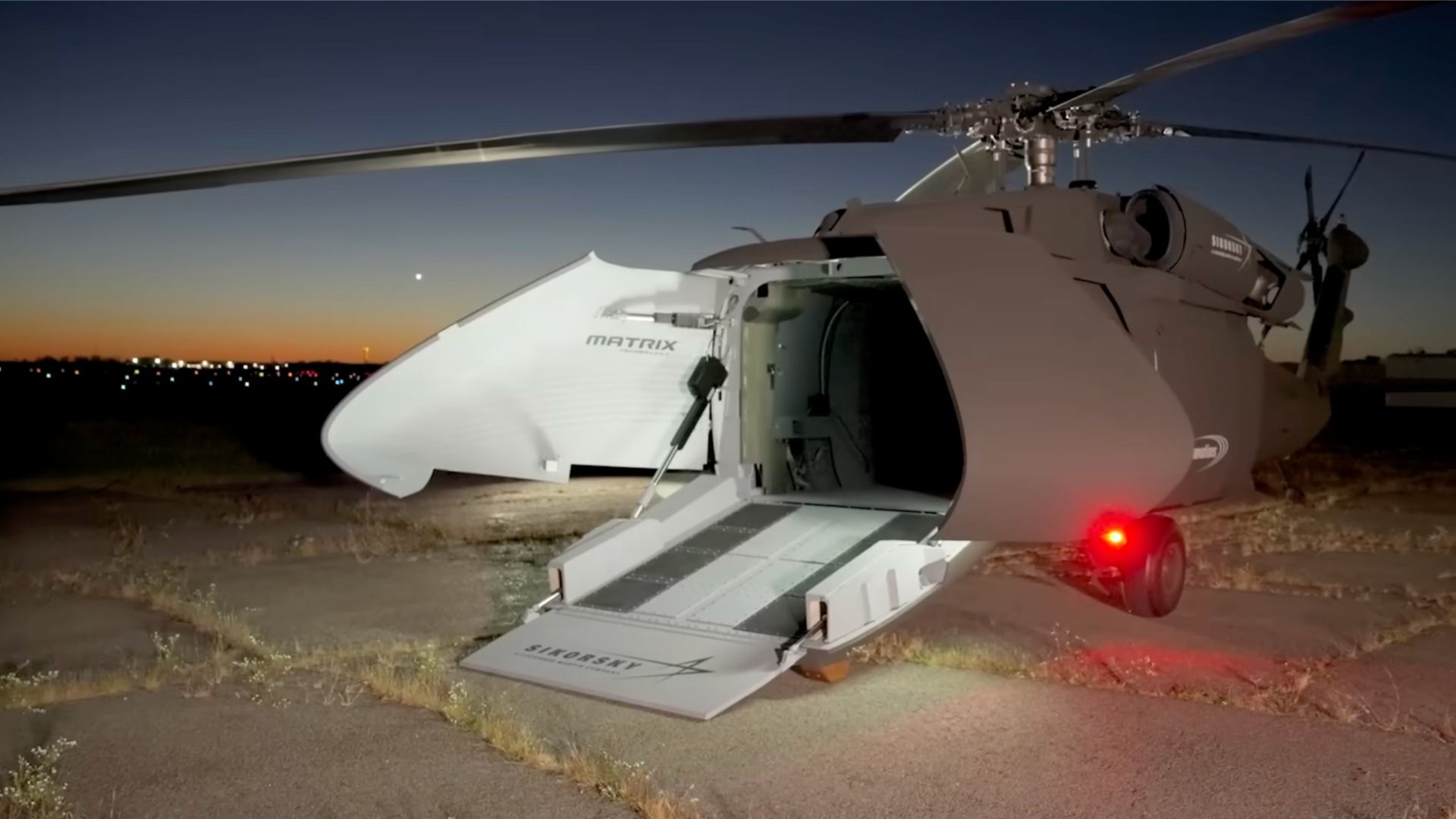 Dusk scene of a brown-gray Sikorsky helicopter on a dirt field with an front door and lowered loading ramp; a red tail light glows as city lights shimmer on the horizon.