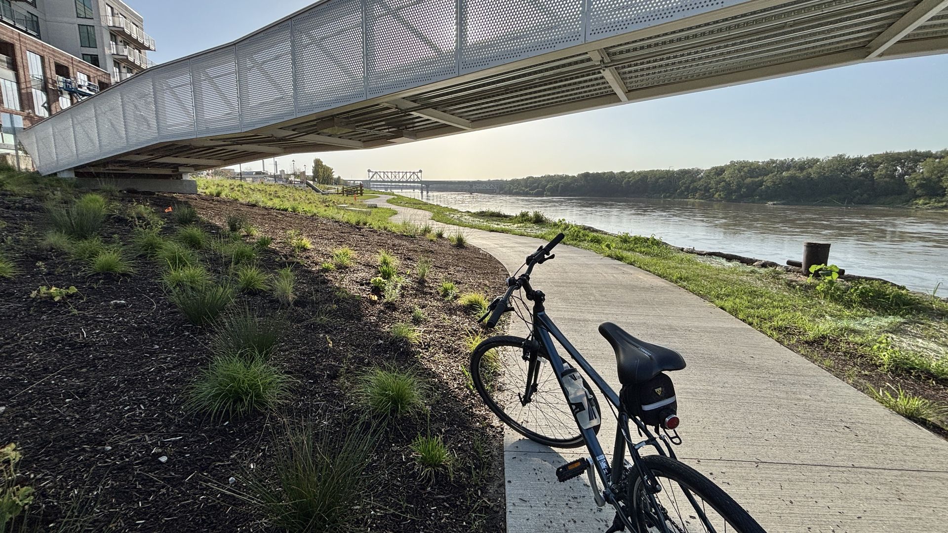 Bicycle on a concrete riverside path under a large perforated metal bridge, with modern brick buildings to the left, a wide river and trees to the right, and a distant bridge across the water.