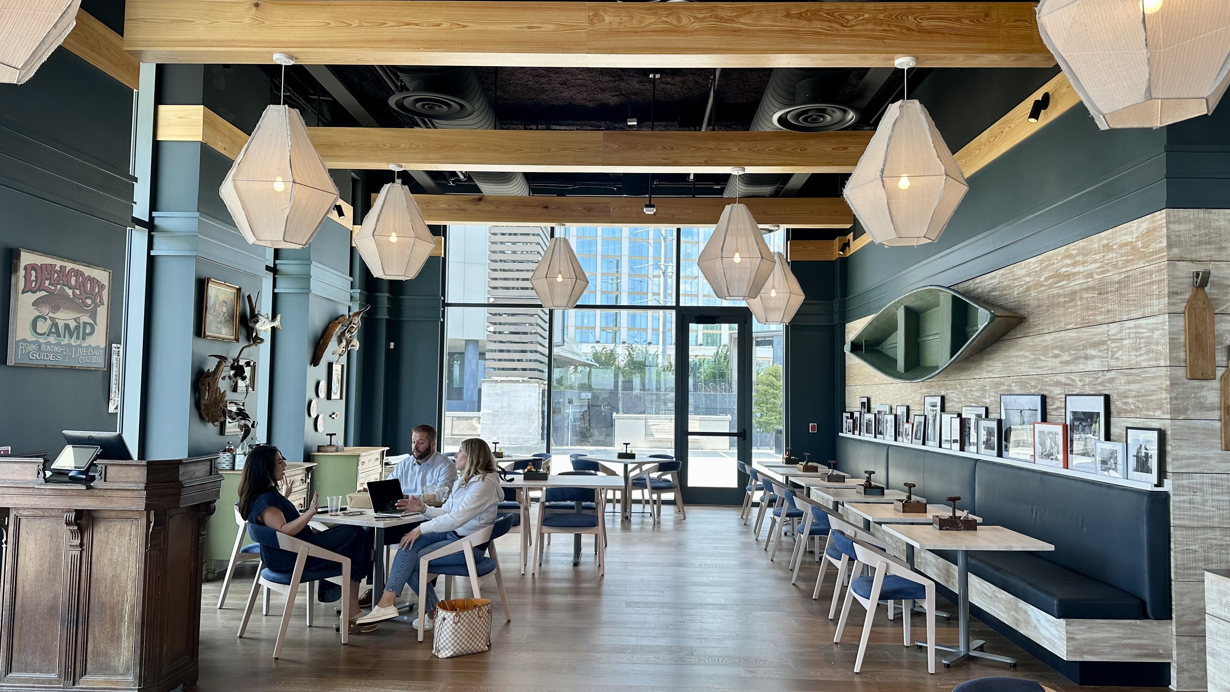 Modern cafe with wooden floors, teal walls, and hanging lantern-style lights. Three people sit at a table near a window with city views, surrounded by framed photos and canoe wall decor.