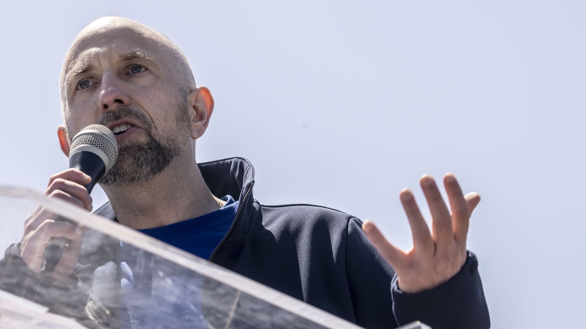 Man with a shaved head and beard speaking into a microphone at a clear podium, wearing a dark jacket and blue shirt, gesturing with his right hand against a pale sky background.
