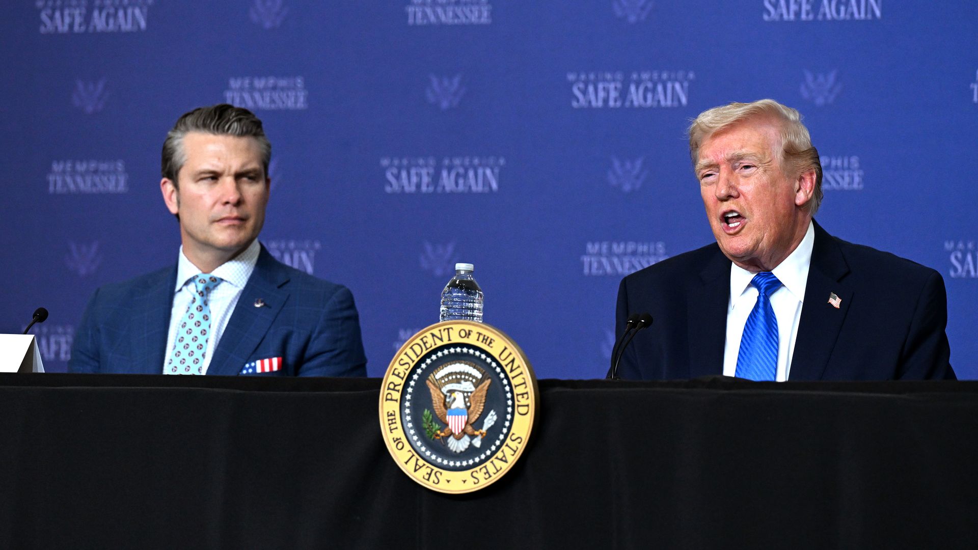 Secretary of Defense Pete Hegseth and President Trump, in suits, at a panel with presidential seal; Trump speaks into a microphone on the right, Hegseth listens. A water bottle sits on the podium; blue backdrop reads Memphis Tennessee and Safe Again.