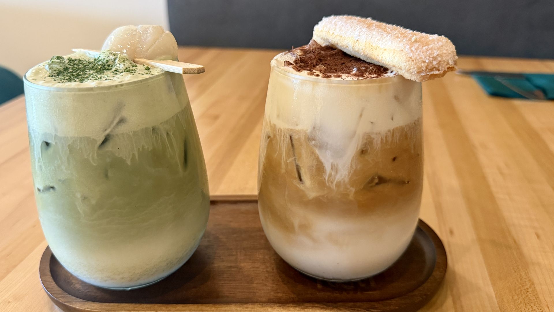Two iced drinks in mason jars on a wooden table: left a green matcha latte with whipped cream and mochi; right a layered iced coffee with cocoa dust and a sugar-dusted pastry on top.