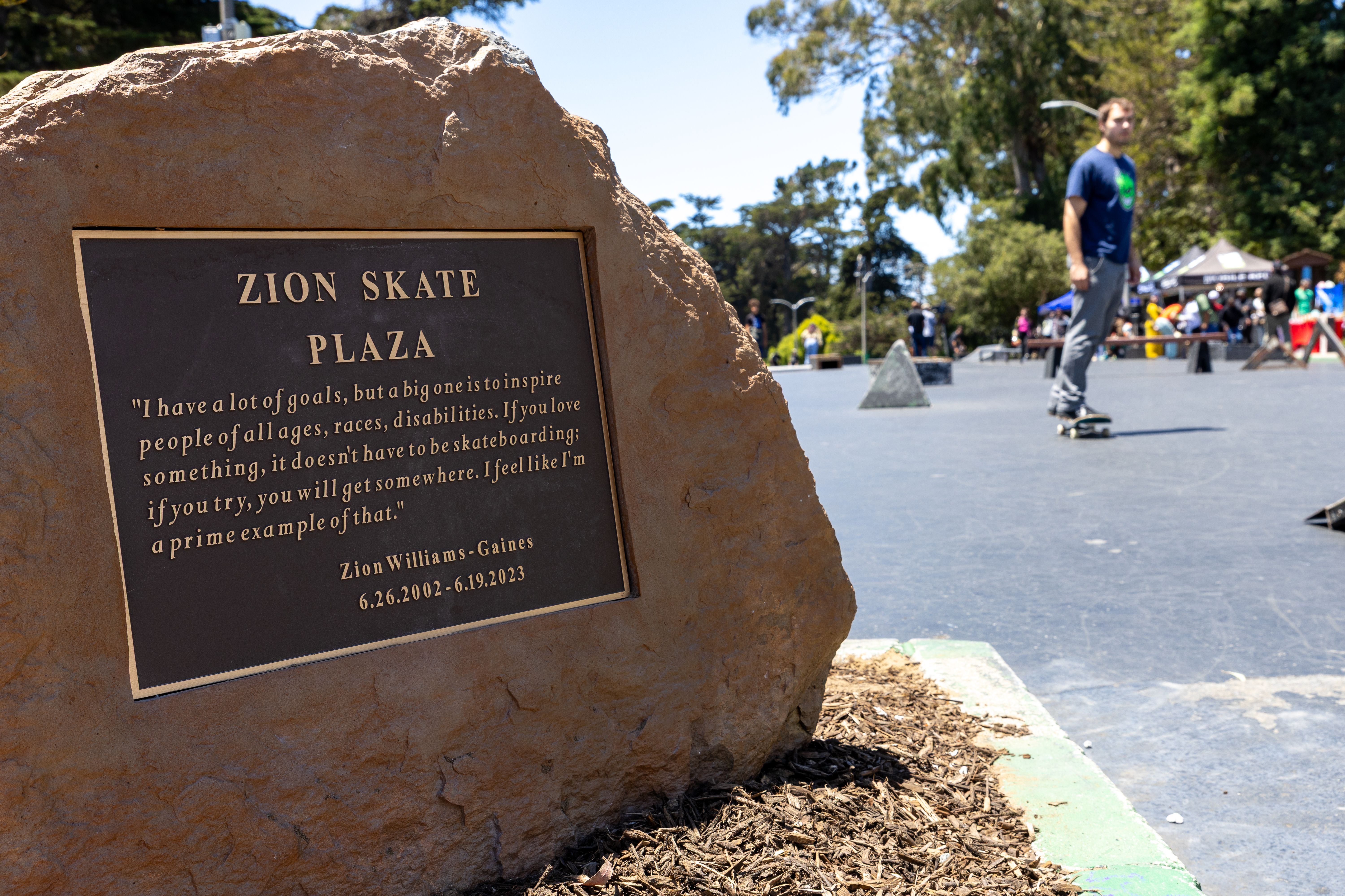 Photo of a plaque that says "Zion Skate Plaza" and skateboarders in the distance