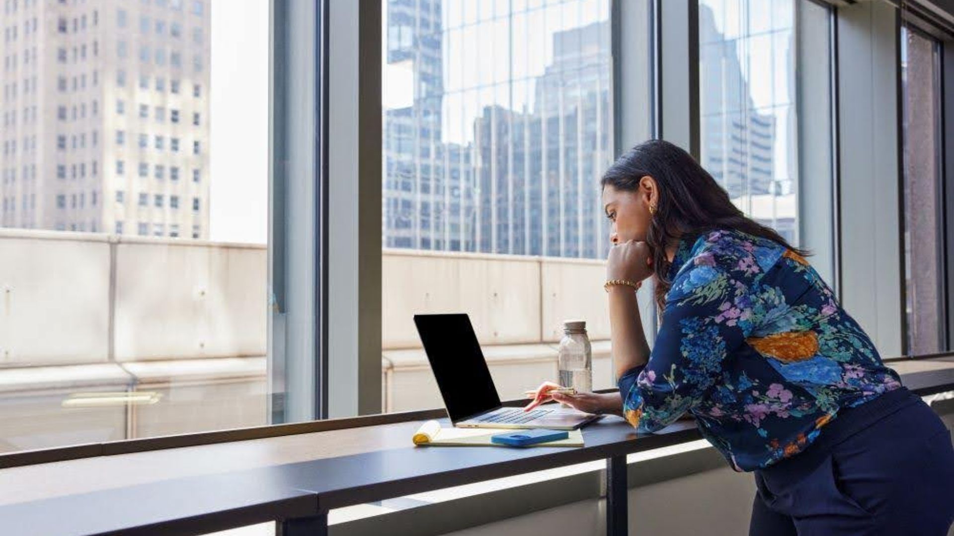 Woman in a floral blouse leaning on a desk, working on a laptop by a large window showing city skyscrapers, with a notebook, phone, and water bottle nearby.
