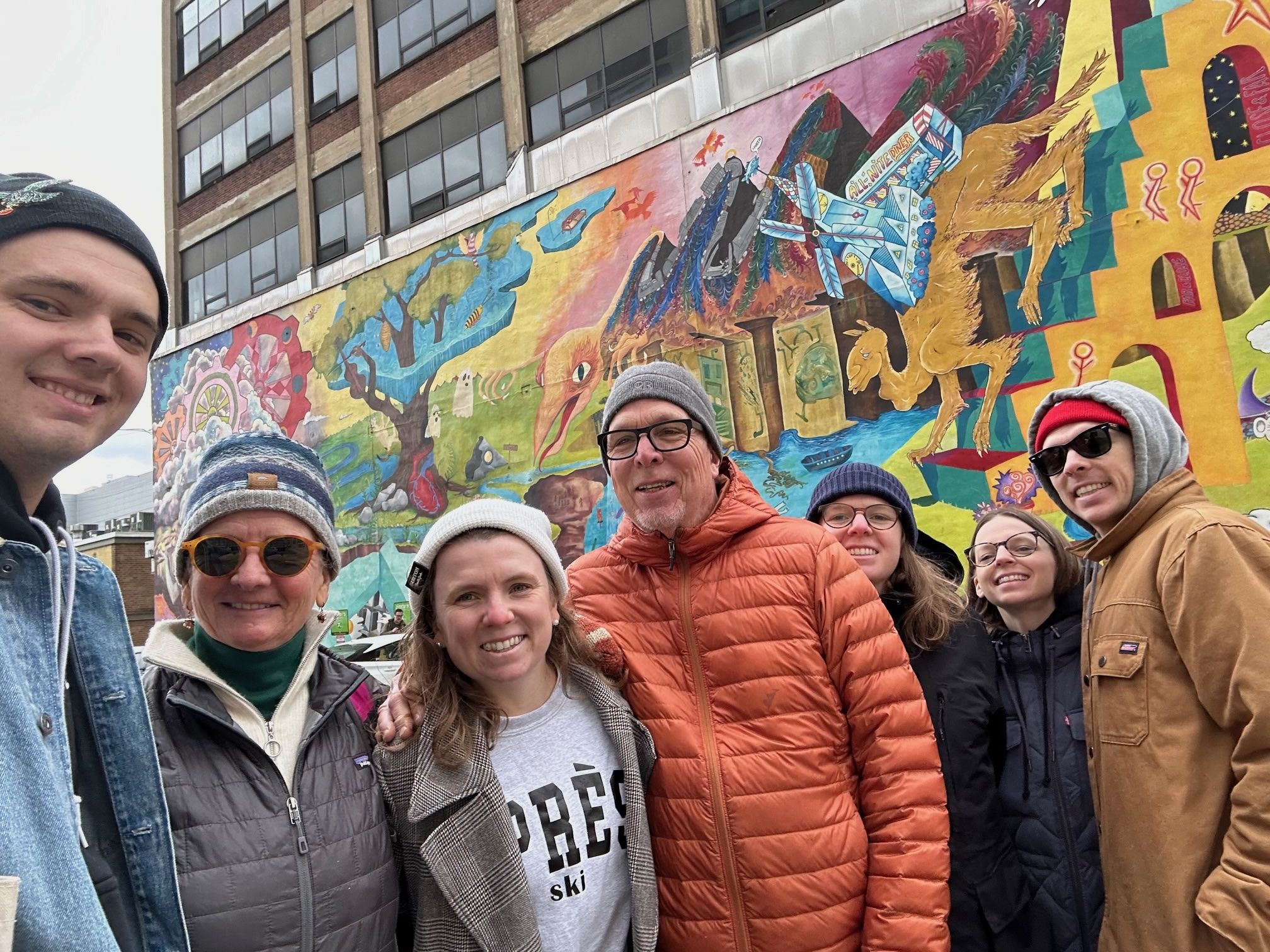 Seven people wearing winter clothes and hats smile in front of a colorful Philly mural.