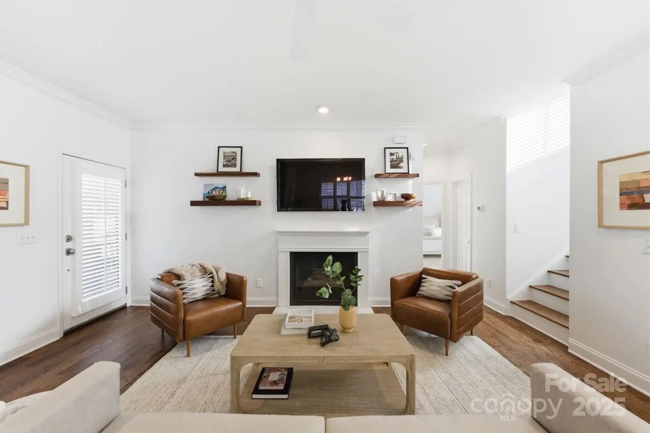 Bright living room with white walls, two brown leather armchairs, beige sofa, wooden coffee table, TV above white fireplace, and floating shelves with decor.
