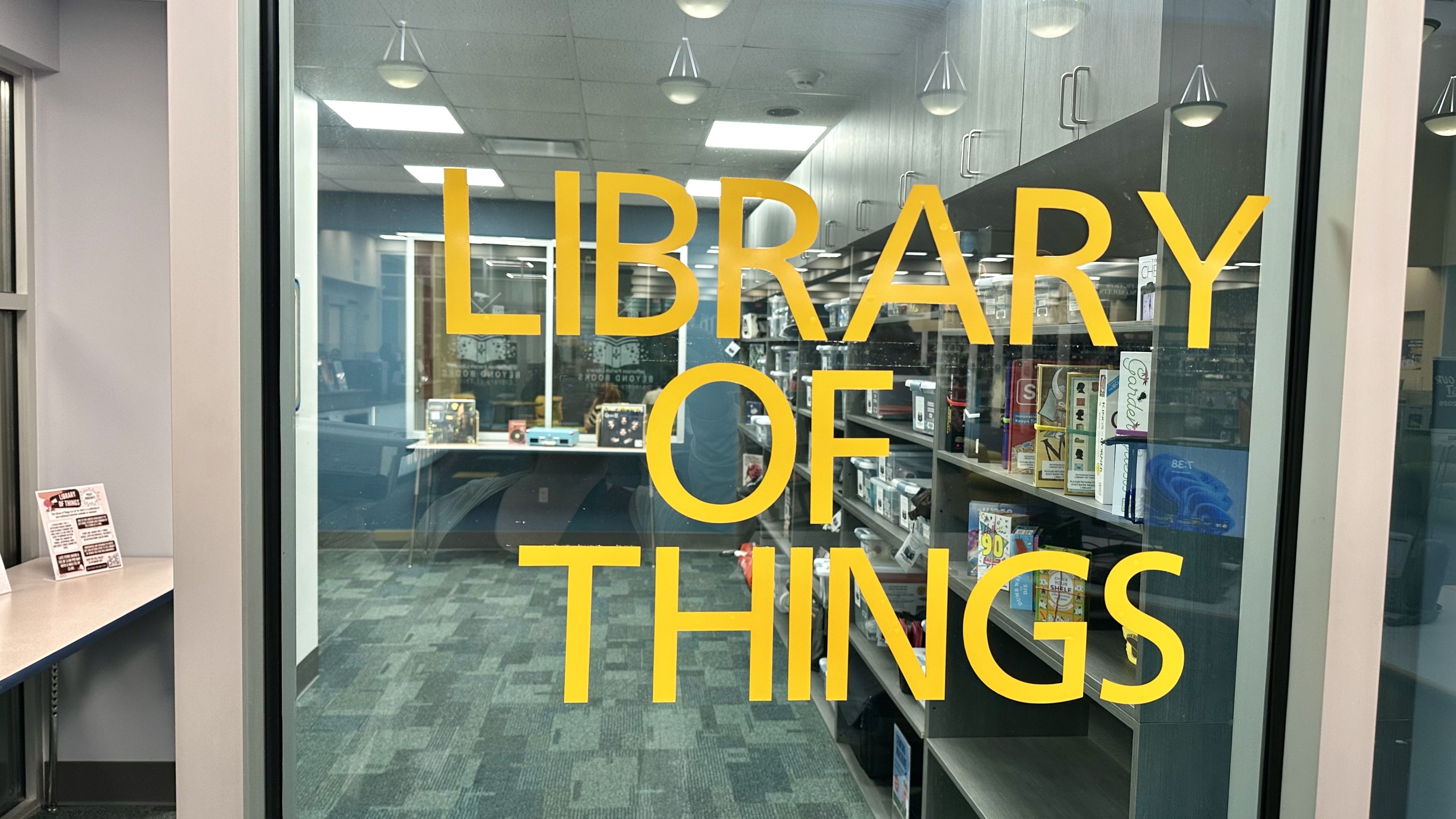 Glass door with large yellow lettering reading "LIBRARY OF THINGS"; inside is a library with shelves, a desk, and a person visible in the background.