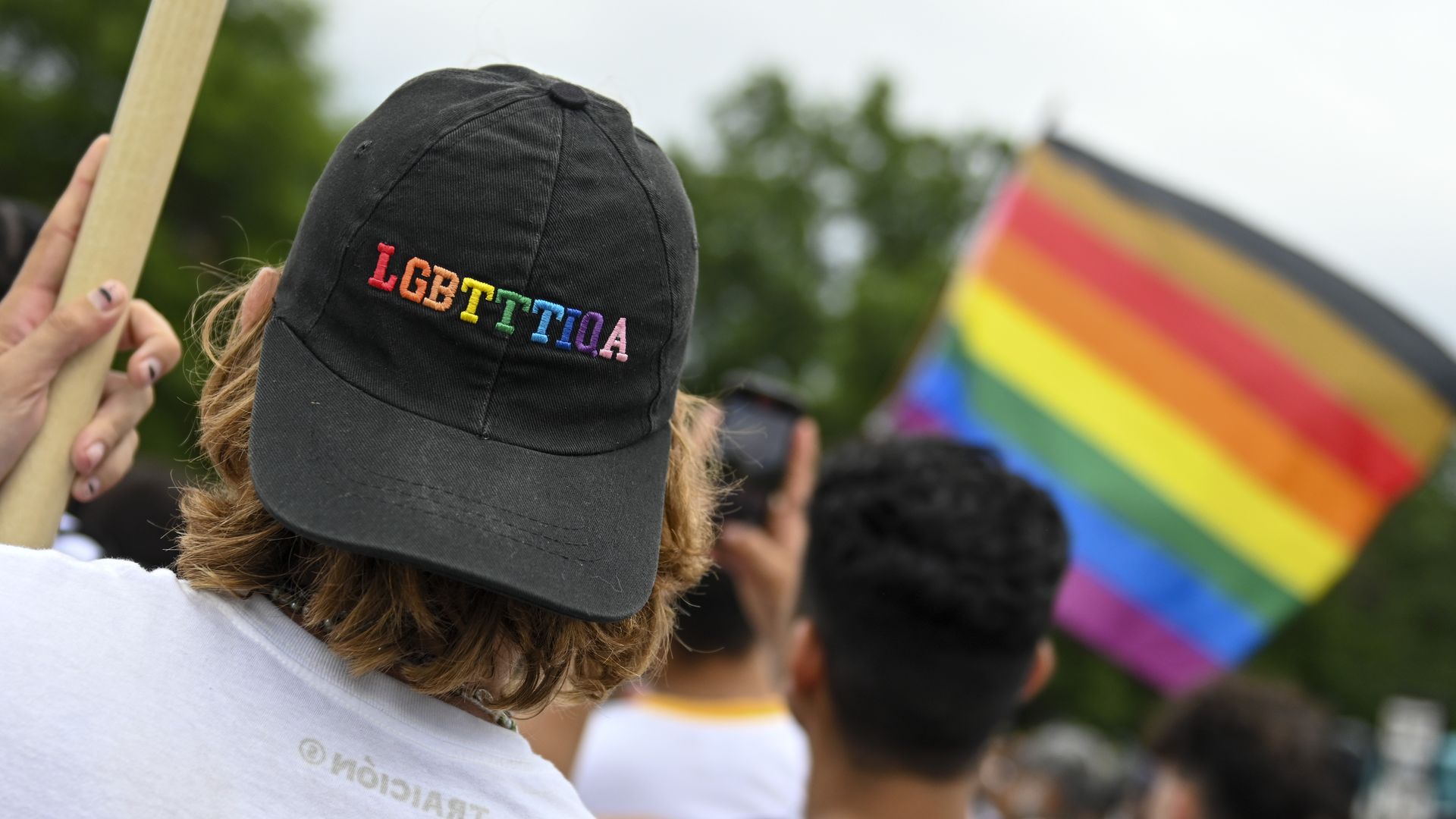 Photo of a person holding a sign facing away from the camera and wearing a cap that says LGBTTTIQA 
