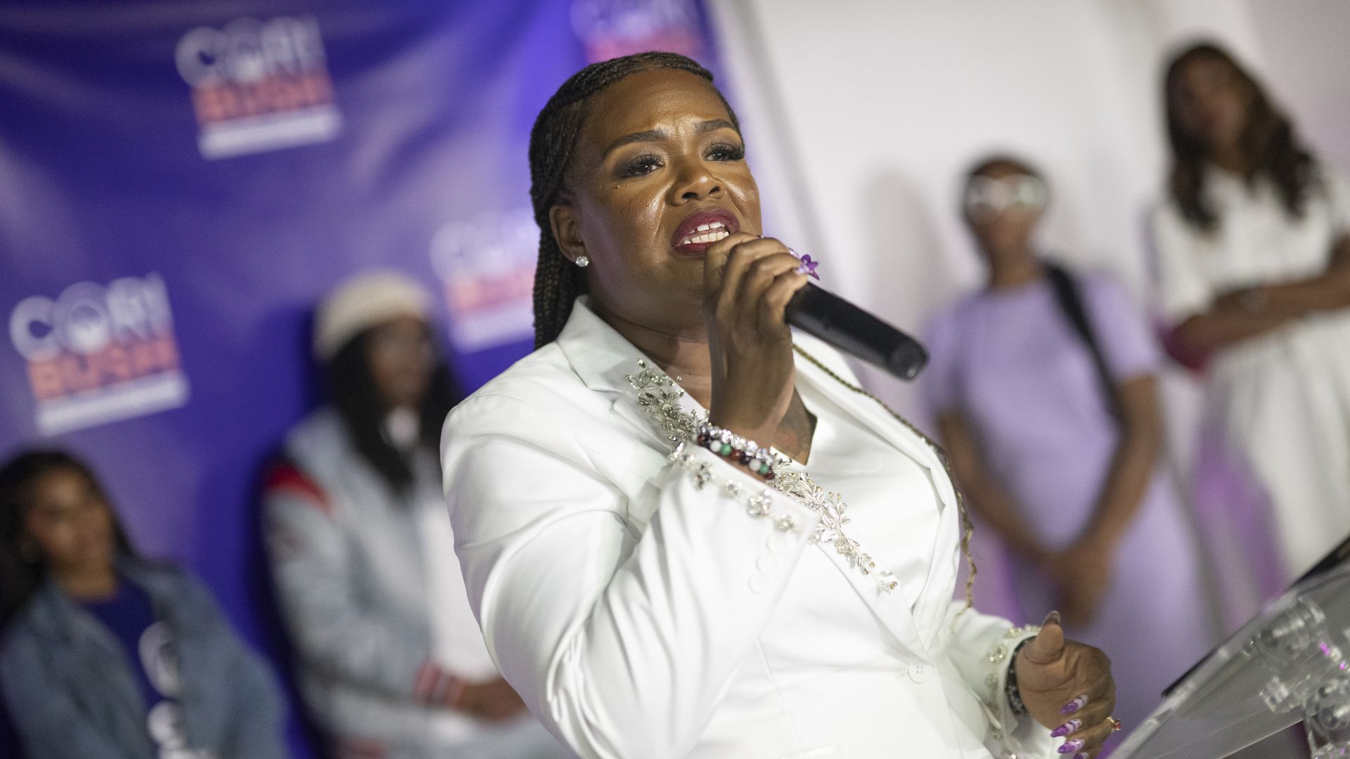 Cori Bush wearing a white suit and speaking into a handheld microphone as she stands behind a glass podium in front of a blue campaign backdrop.