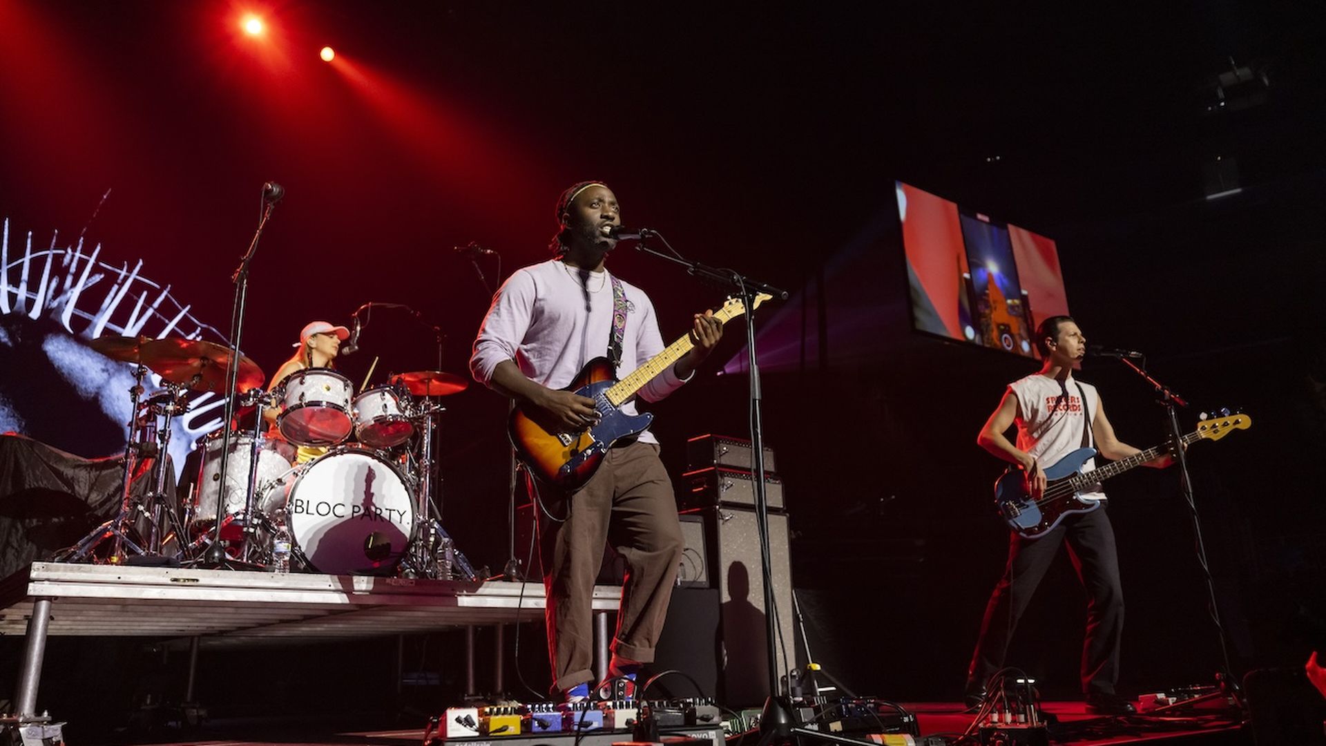 A photo of a band on stage, the lead holding an electric guitar and singing into a microphone with red lights overhead.