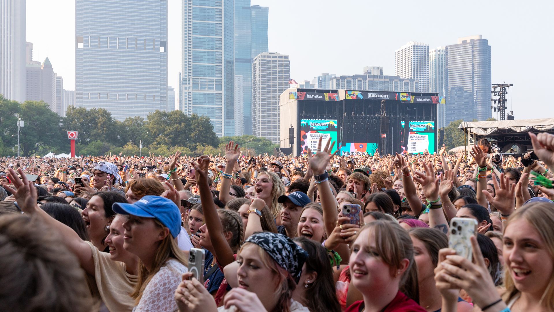 Massive outdoor concert crowd with hands raised, a large stage with colorful screens, and a city skyline of tall glass buildings in the background.