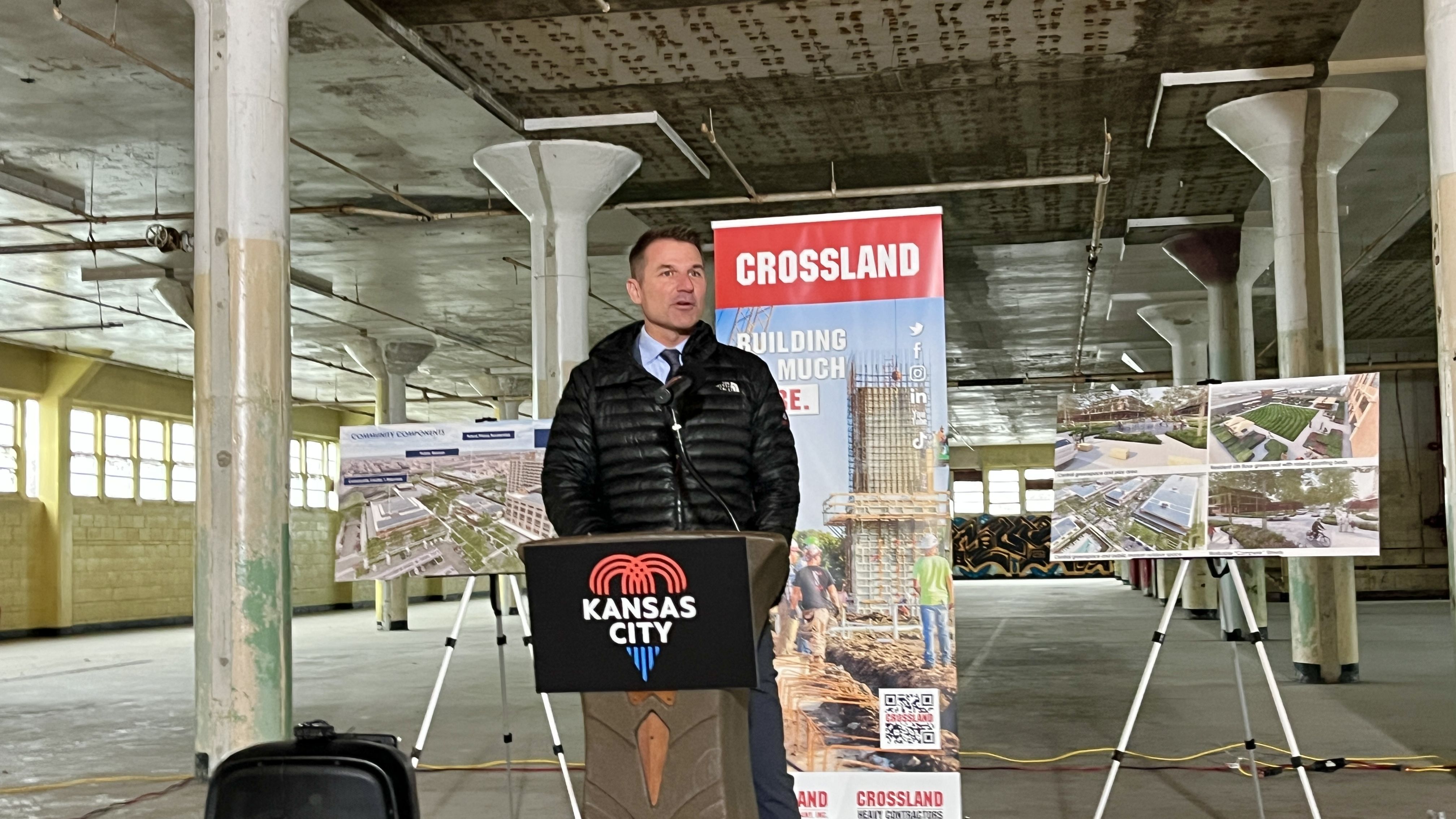 Jonathan Arnold in black jacket at Kansas City podium inside a large industrial space, speaking with Crossland construction banners and building project boards behind him.