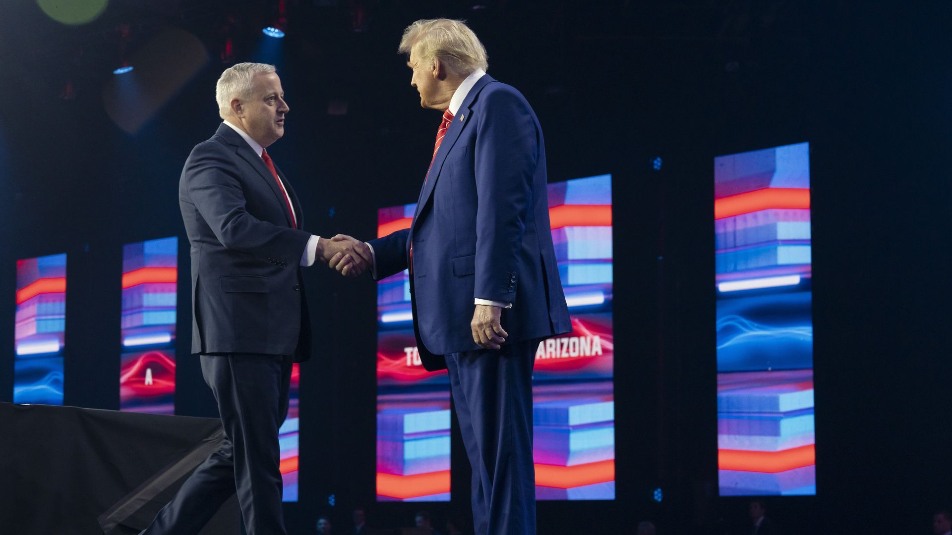 Two men in suits shaking hands on a stage with blue, red, and white illuminated panels in the background.