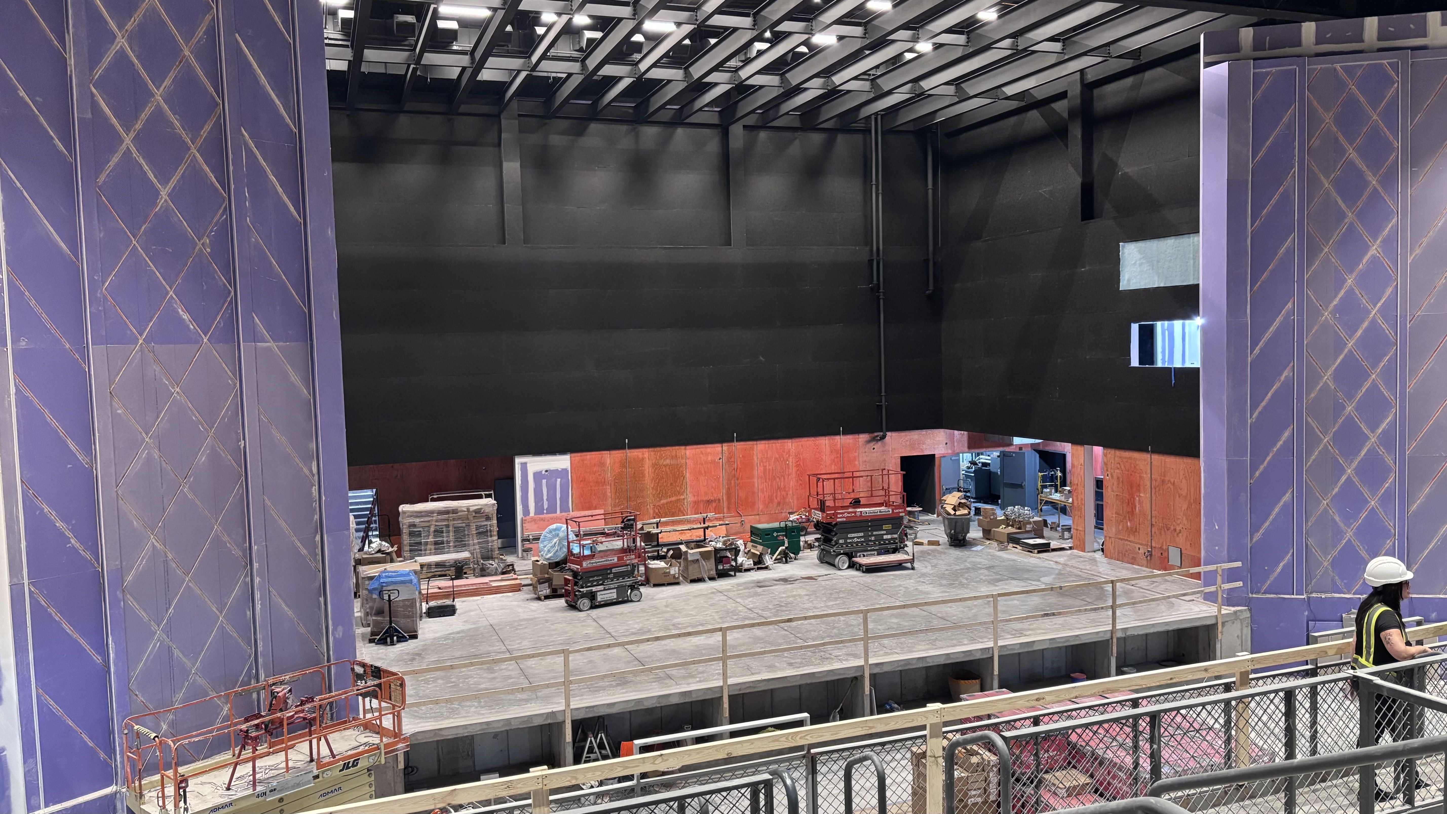 Inside a theater under renovation: purple columns with diamond patterns frame a black back wall; visible girders and lights; construction equipment and a worker in a yellow vest on site.