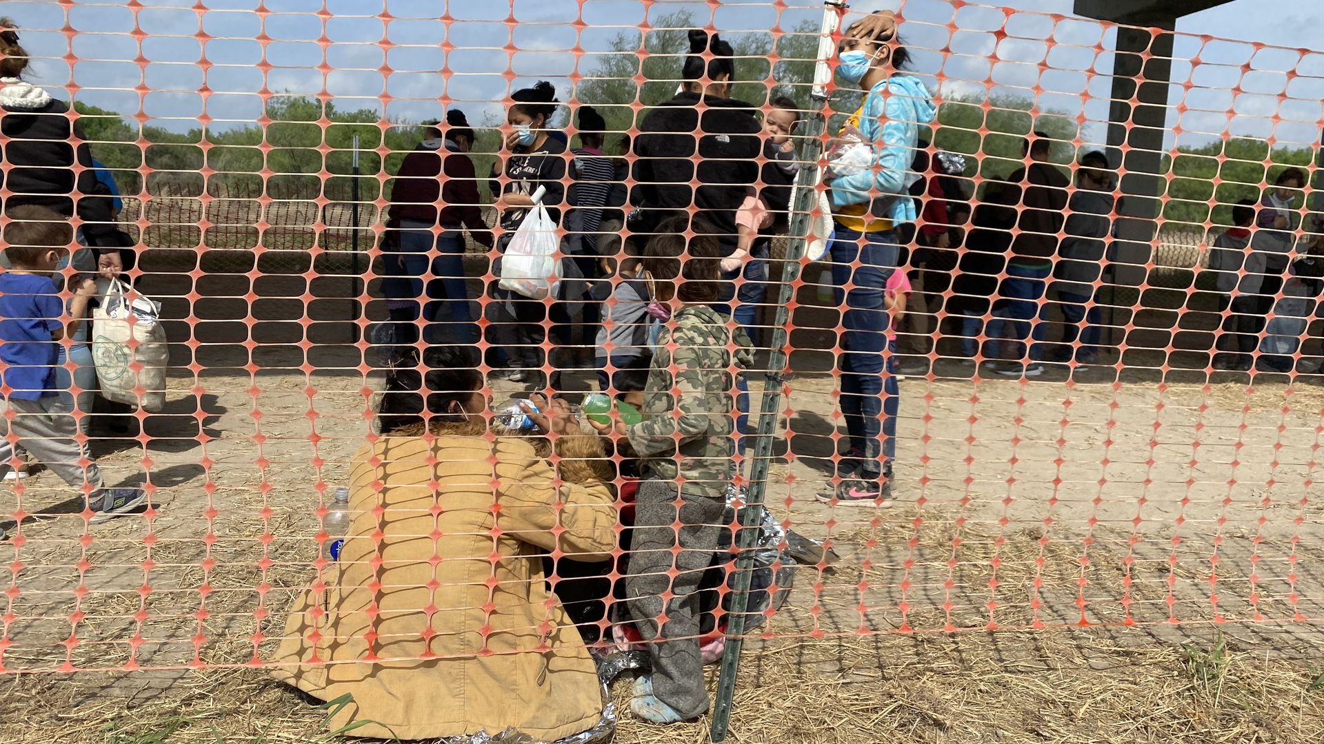 Migrant mothers and young children sit on the ground outside behind orange safety netting. 