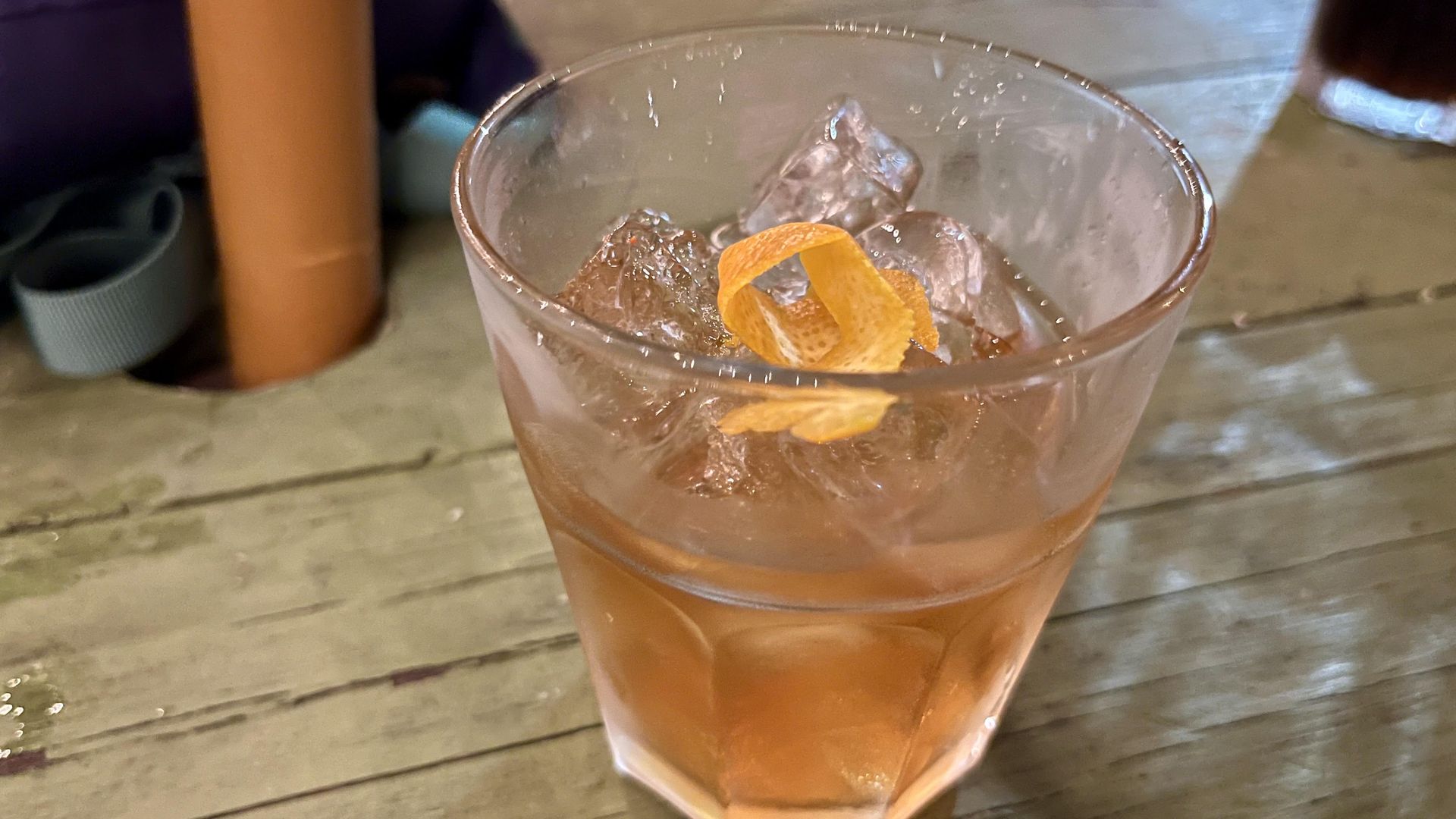 A close-up of an old fashioned cocktail in a multi-faceted glass with ice and a curled orange peel garnish, set on a weathered wooden picnic table at Slow Ride in San Antonio.