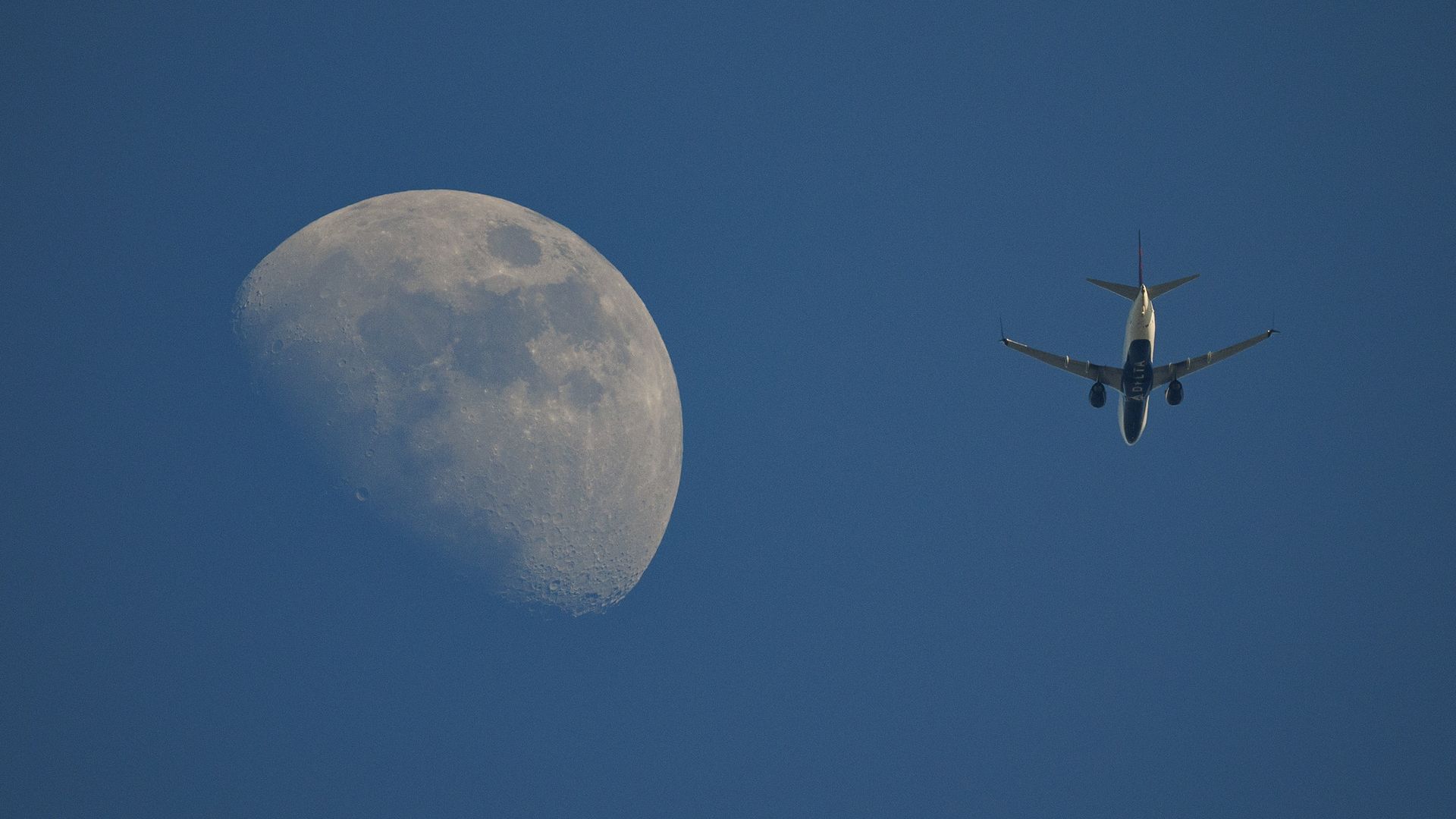Airplane flying in a clear blue sky near a large, detailed half moon visible in daylight.