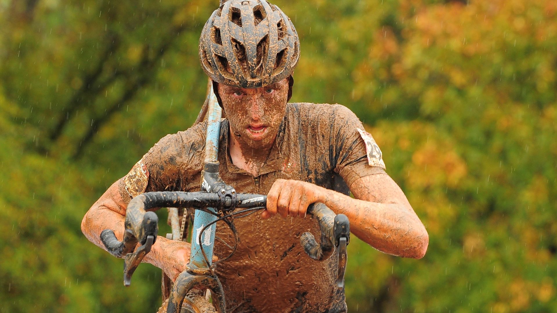 A determined-looking man covered in mud and wearing a bike helmet is carrying a bike on his shoulder.