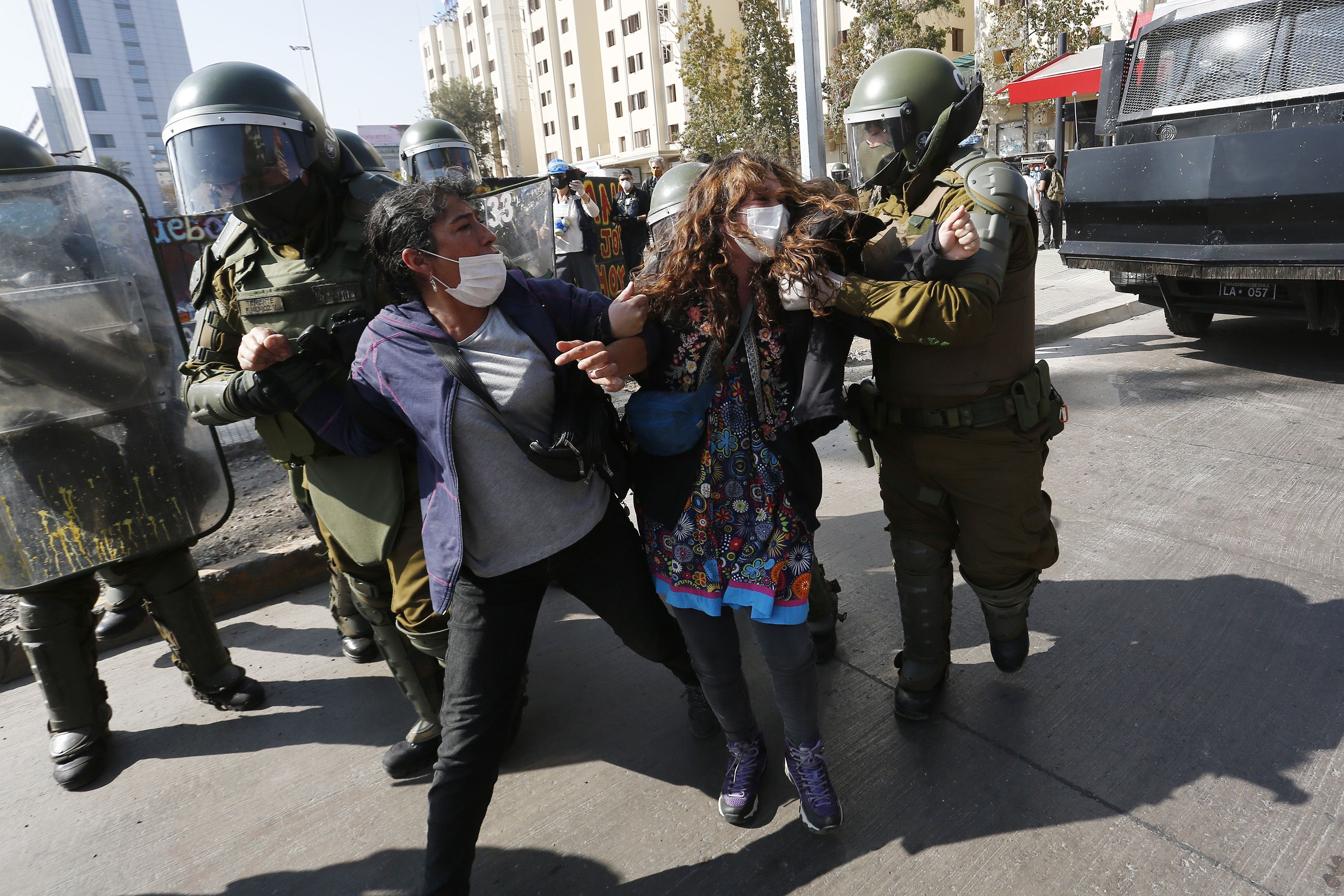 In this image, a masked woman and a masked man are handled by police wearing armor