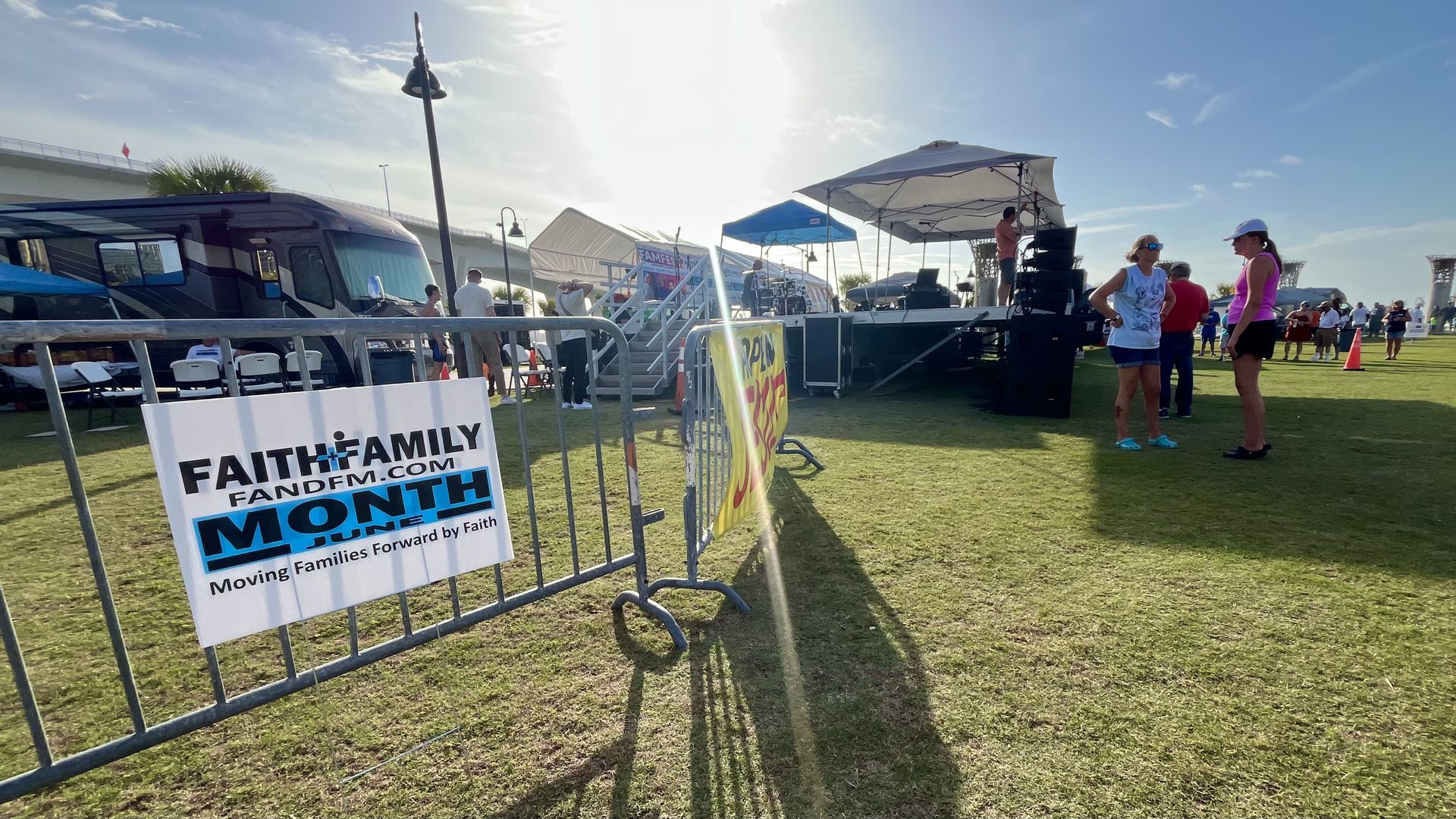 A sign on a barricade that reads "FIATH AND FAMILY MONTH fandfm.com Moving Families Forward by Faith." In the background, people mill around a stage set up on a lawn.