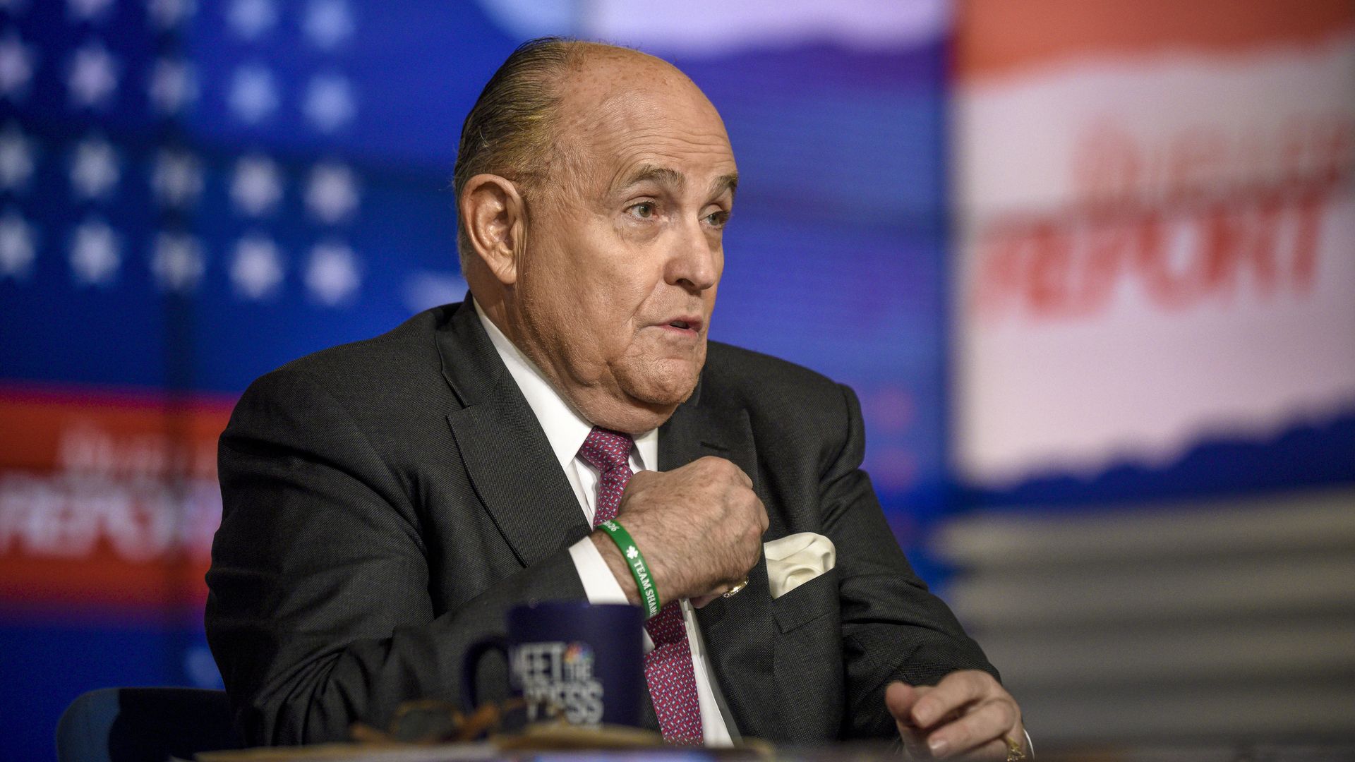 In this image, Rudy sits in a suit and tie and gestures to himself while sitting at a desk with the American flag in front of him.