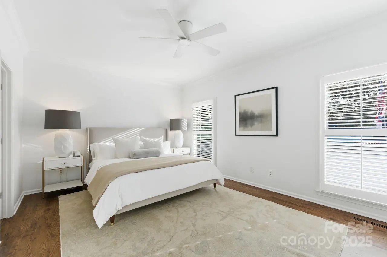 Bright bedroom with white walls, ceiling fan, large beige rug, wooden floor, beige bed with white and beige pillows, two lamps with dark shades, and a framed abstract picture on wall.