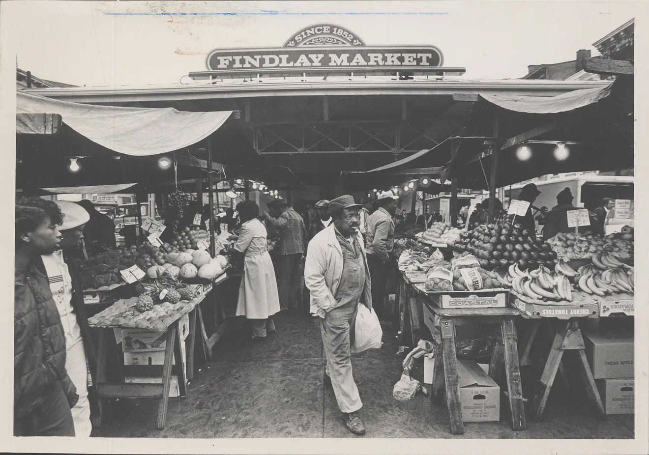 Black-and-white photo at Findlay Market, a bustling open-air fruit market with produce on wooden tables; shoppers browse as a man in a cap stands center foreground.