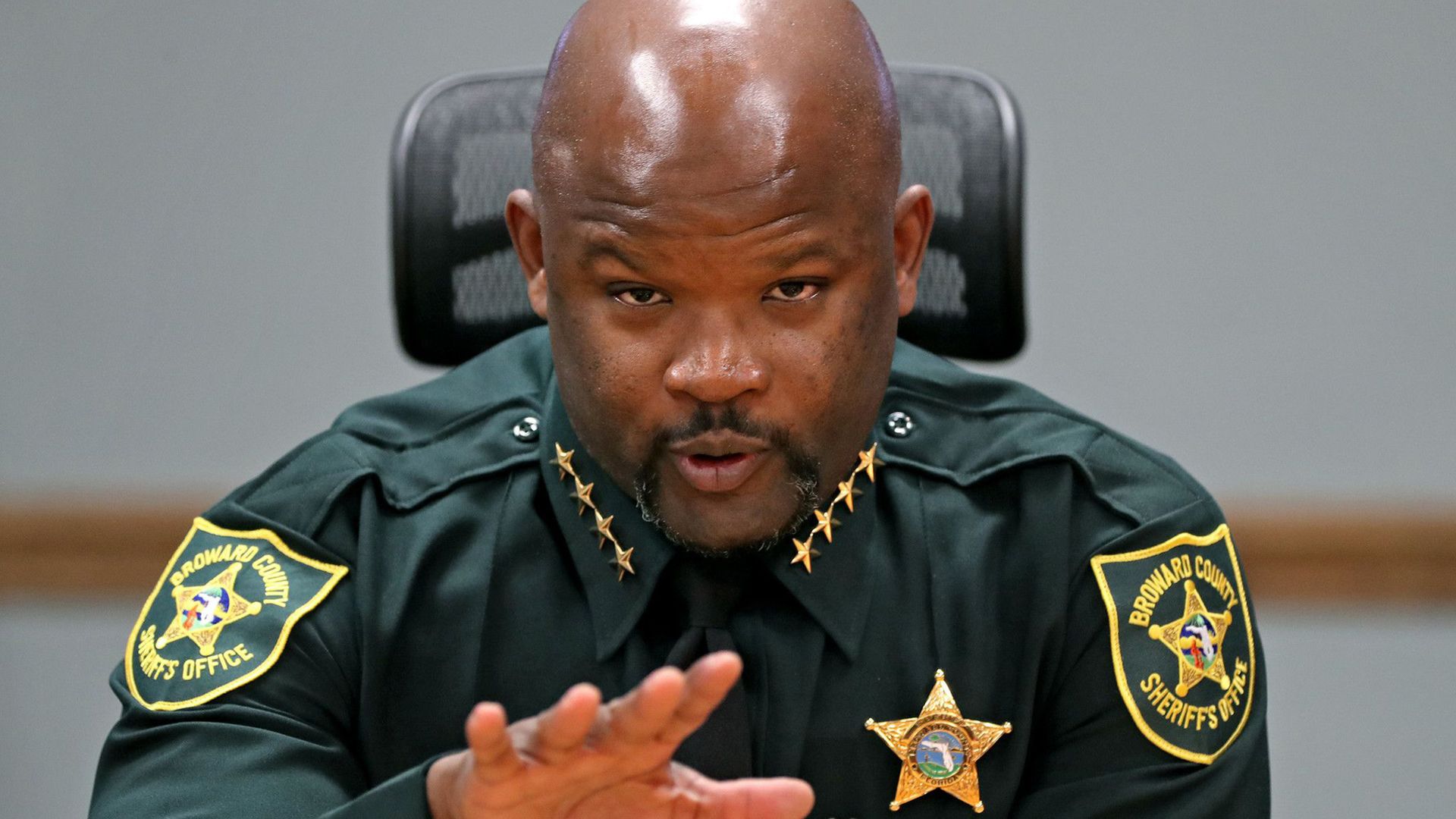 Sheriff Gregory Tony sits in a chair wearing his green police uniform with his right arm stretched out pointed towards the camera. His chin is slightly faced down.