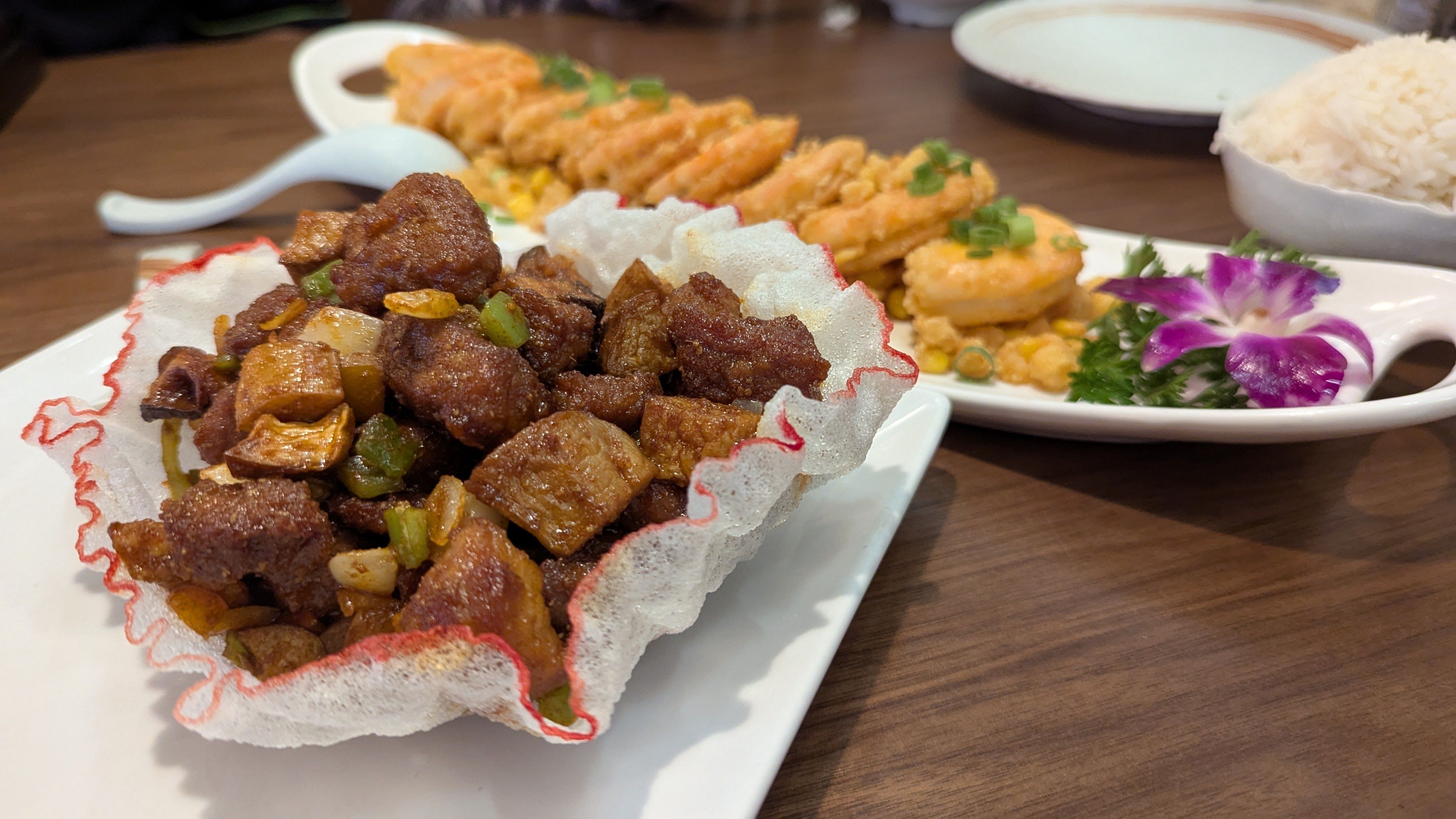 Close-up of a dish with crispy fried meat cubes, onions, and green peppers served in a white, red-tinted crispy shell on a white plate, with fried shrimp and white rice in background.