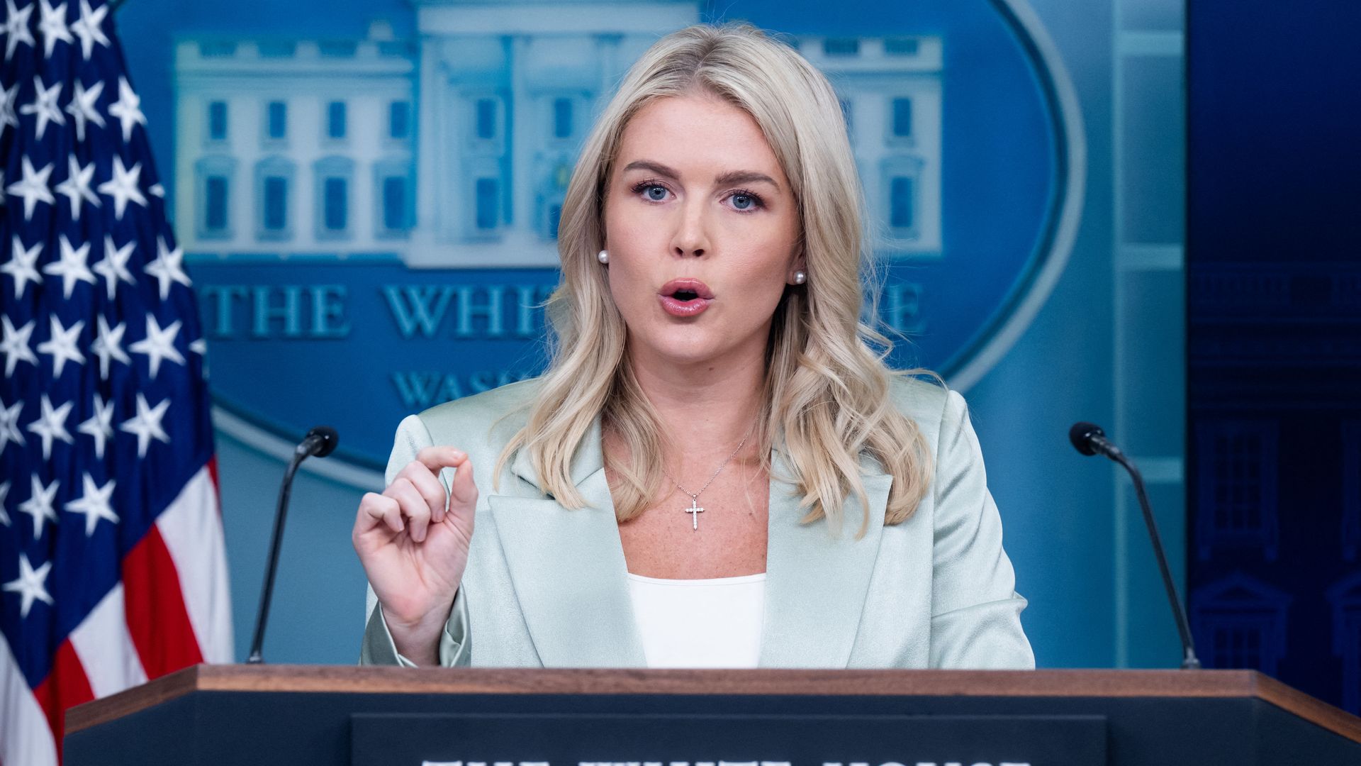 Karoline Leavitt, wearing a light green silky blazer and a white shirt, gestures as she speaks from behind a podium.