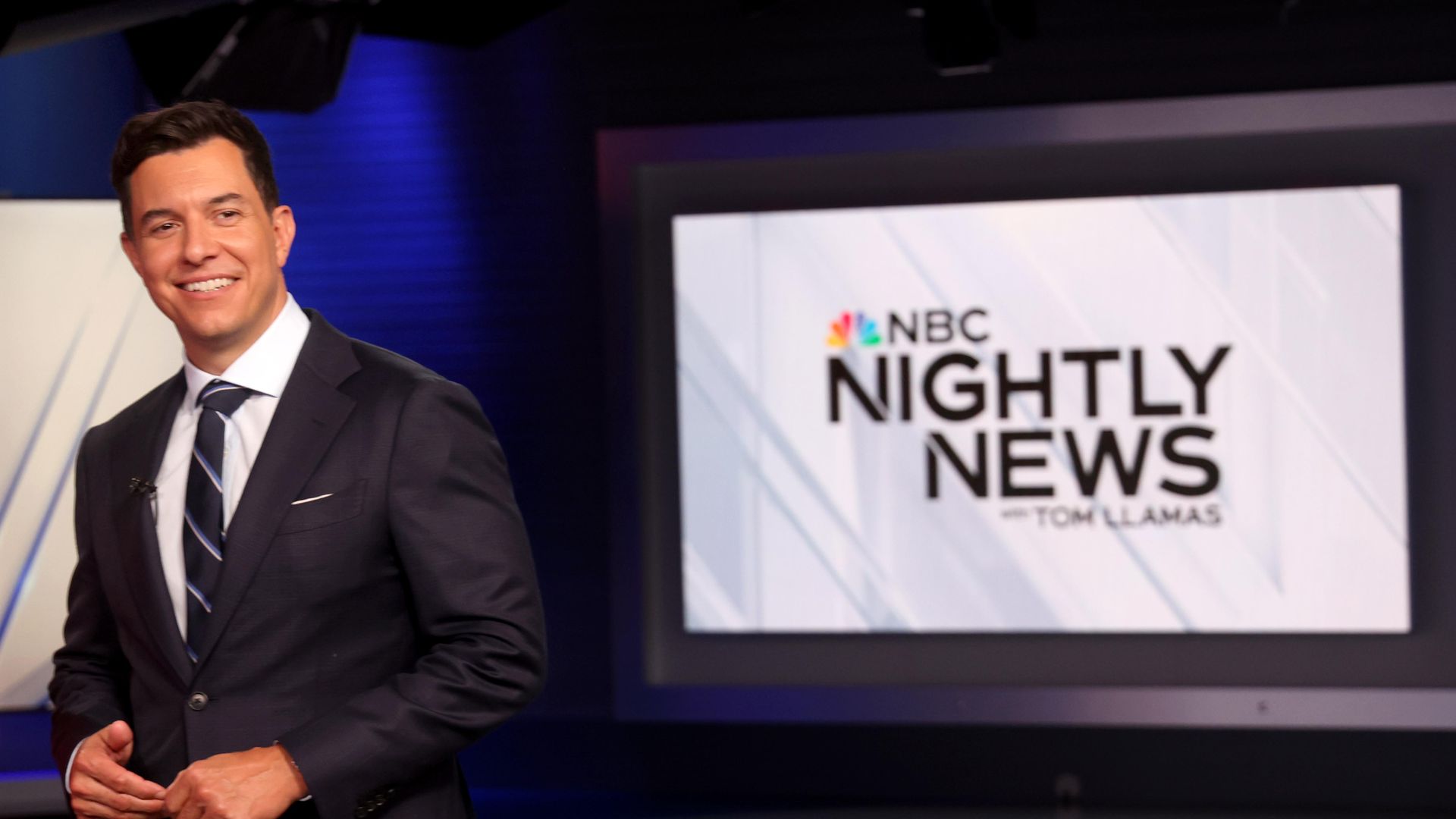 Smiling male news anchor in a dark suit and striped tie standing in a studio with a screen showing "NBC Nightly News with Tom Llamas" logo in the background.
