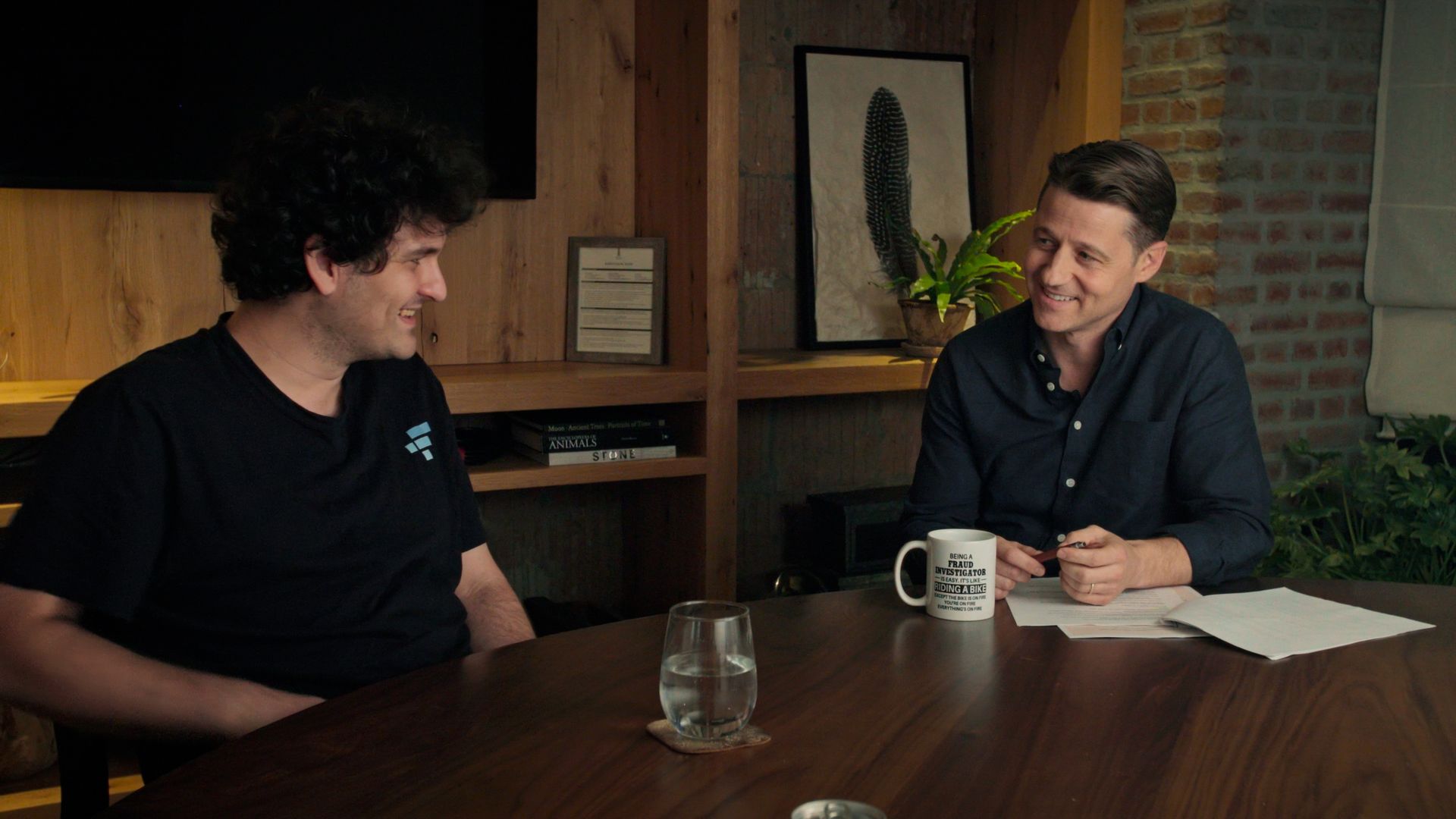 Two men sit at a wooden table in a cozy studio with brick wall and shelves. Left: curly-haired man in a black T-shirt; right: man in a dark blue shirt smiles. Papers and a mug sit between them.