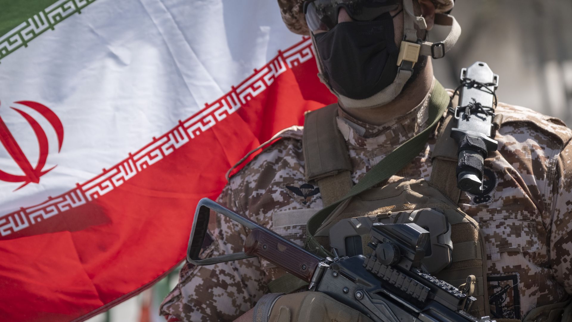 A person wearing a camouflage military uniform and a black mask while holding a gun stands in front of an Iranian flag.