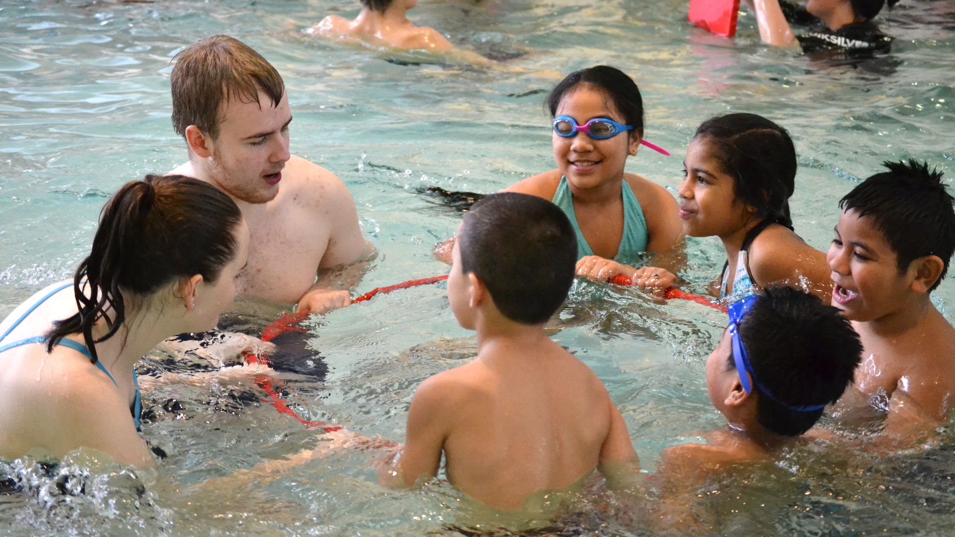 Children in a swimming pool with an instructor all holding onto a hula hoop.