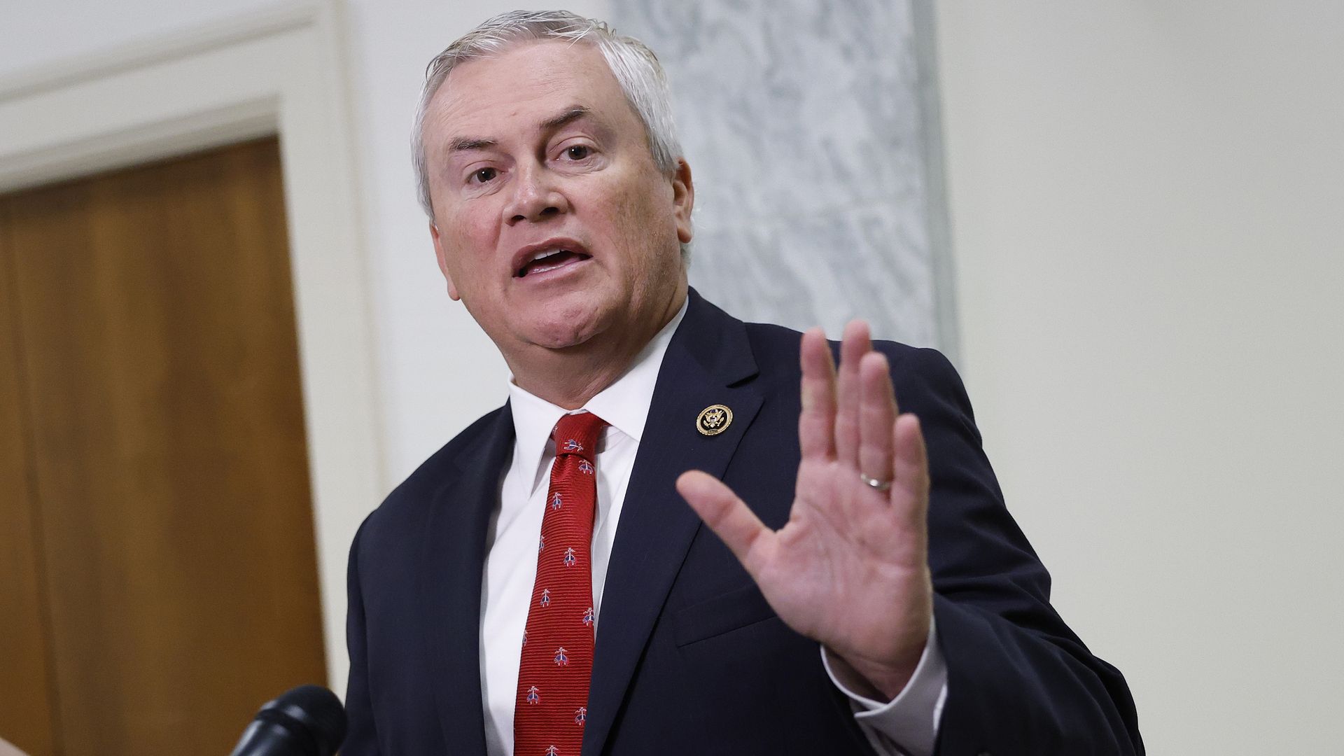 White-haired man in a navy suit, white shirt, and red tie speaking and raising his hand near a microphone, with a wooden door and marble wall background.