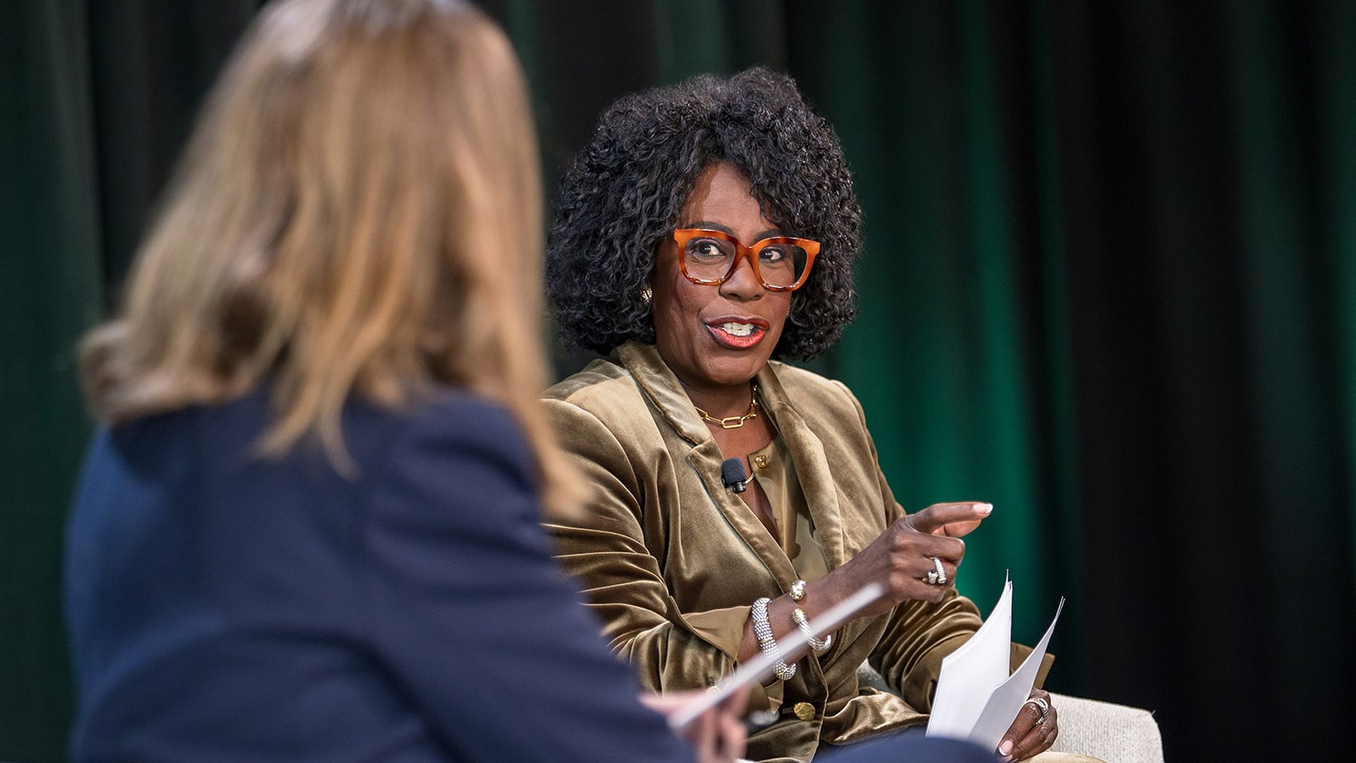 Black woman with curly hair and orange glasses wearing a gold velvet suit speaks in a panel discussion, holding papers; another person with their back to the camera sits nearby.