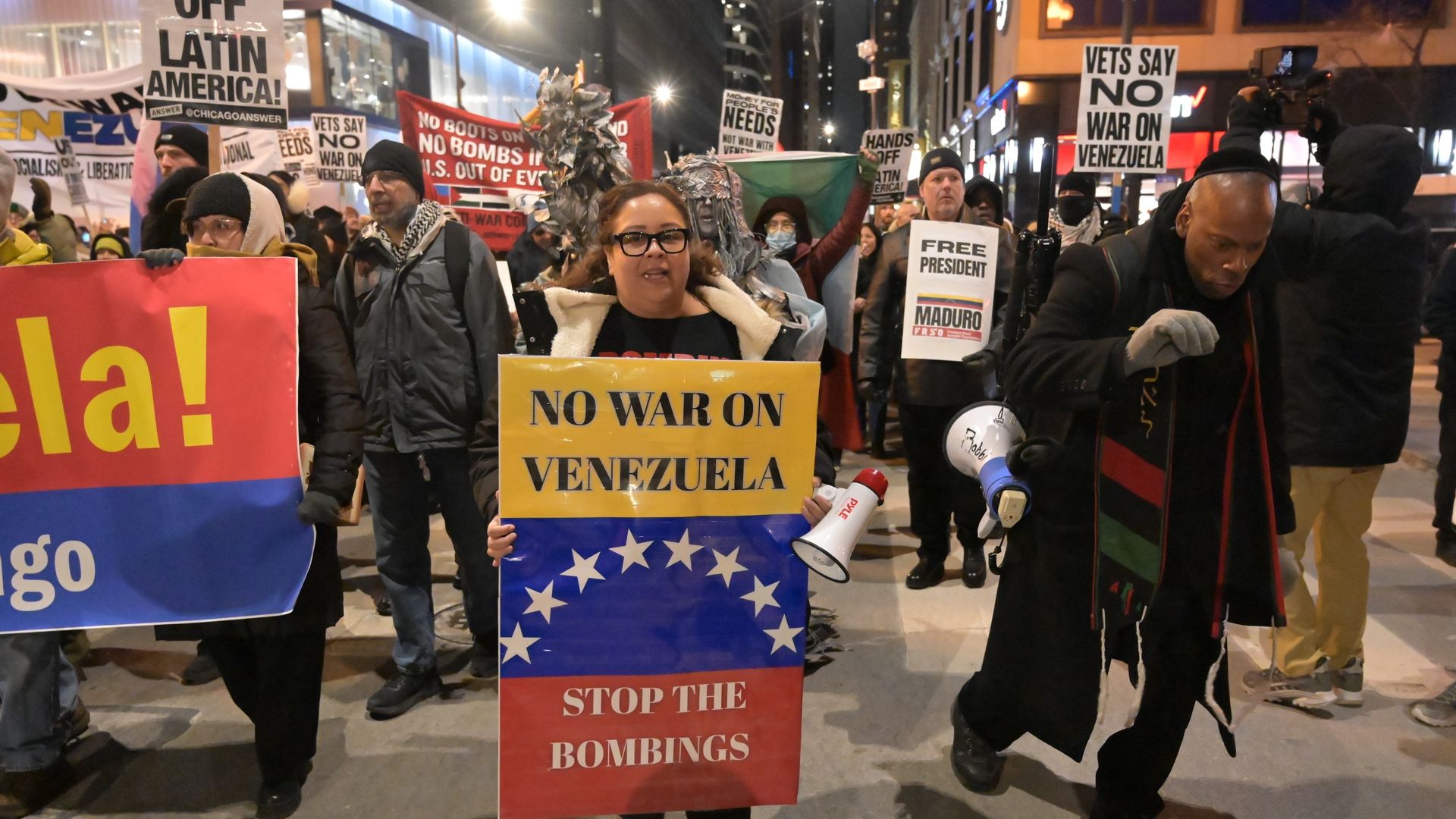 Protesters at night hold signs reading "No war on Venezuela" and "Stop the bombings," with others carrying signs opposing war on Venezuela and supporting President Maduro.