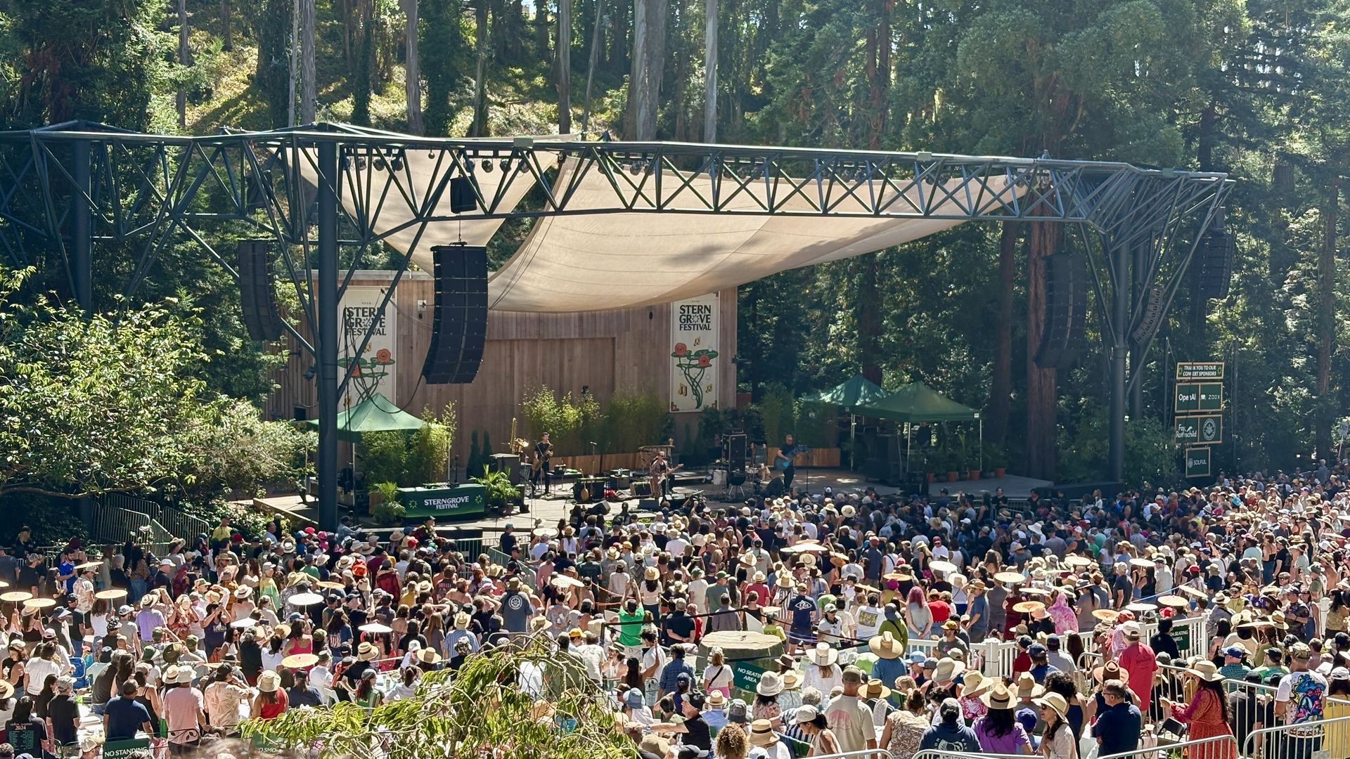 Outdoor concert at Stern Grove Festival. A large metal truss stage with a beige canopy in a forest; a crowded audience in hats watches the band.