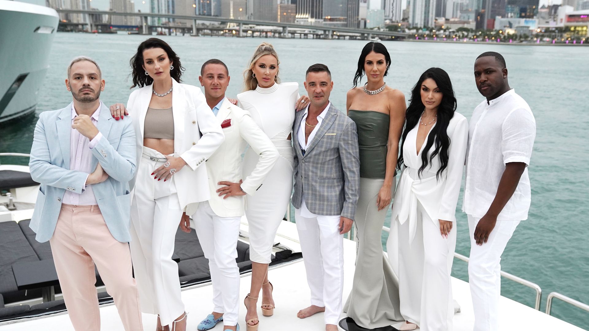 A group of people in white and light-colored suits stands on the deck of a yacht 