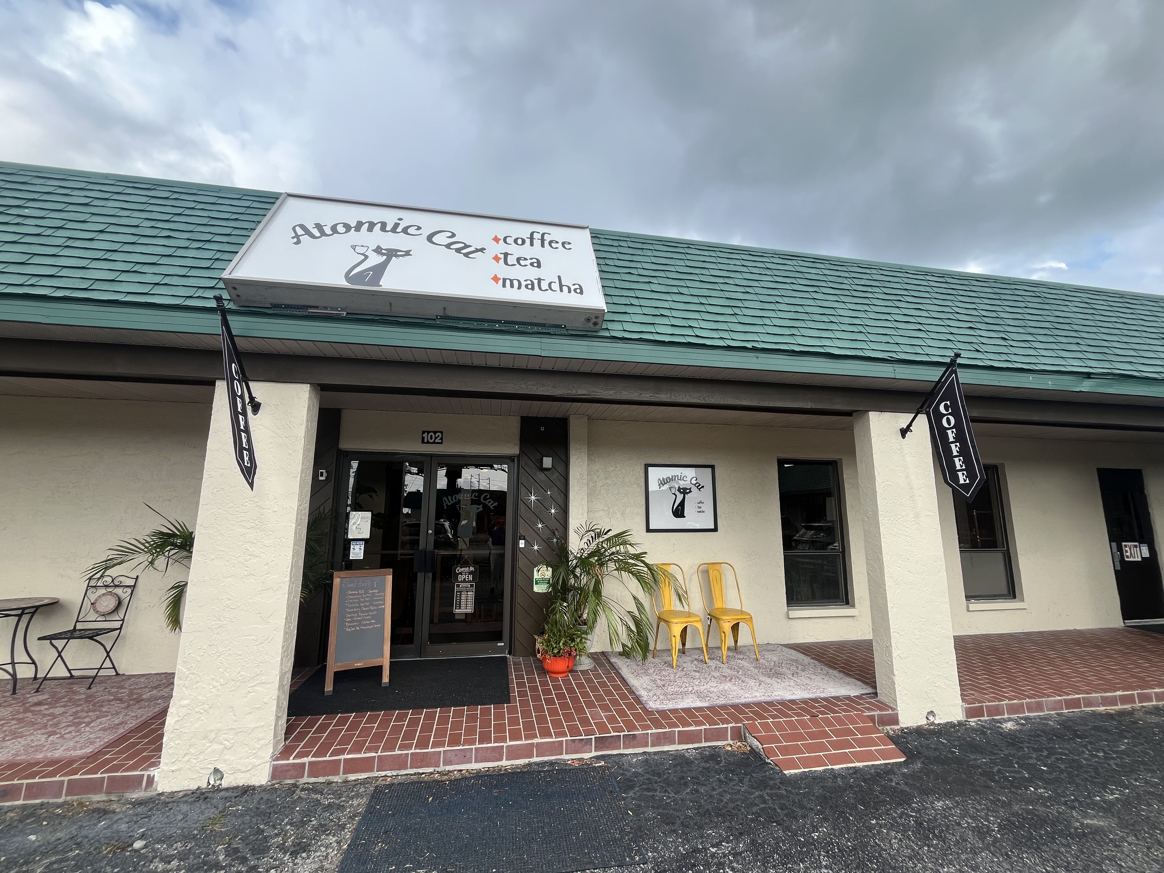 Front of Atomic Cat cafe with a white sign showing a cat silhouette and the name "Atomic Cat". A vertical list reads coffee, tea, matcha. Teal roof, cloudy sky, glass doors, yellow chairs outside.