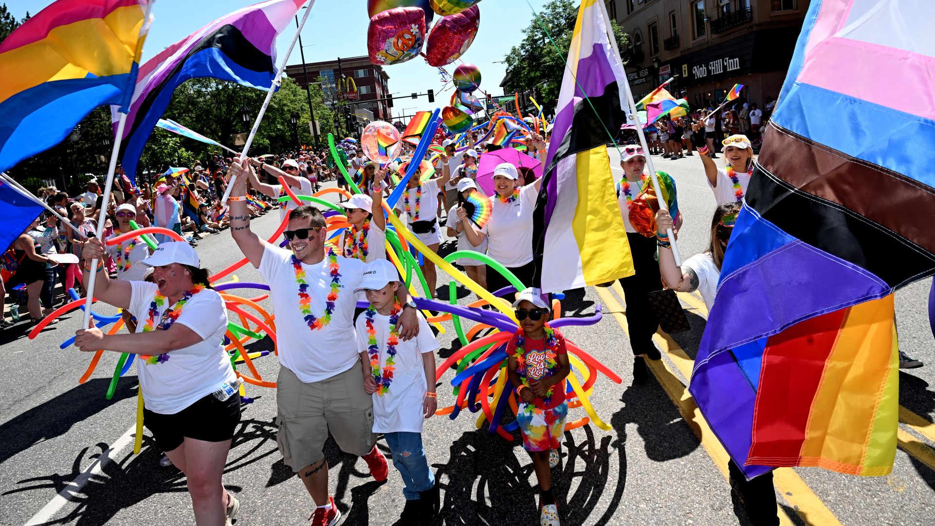 Several people wave colorful flags during a parade procession with rainbow-colored flags and balloons.
