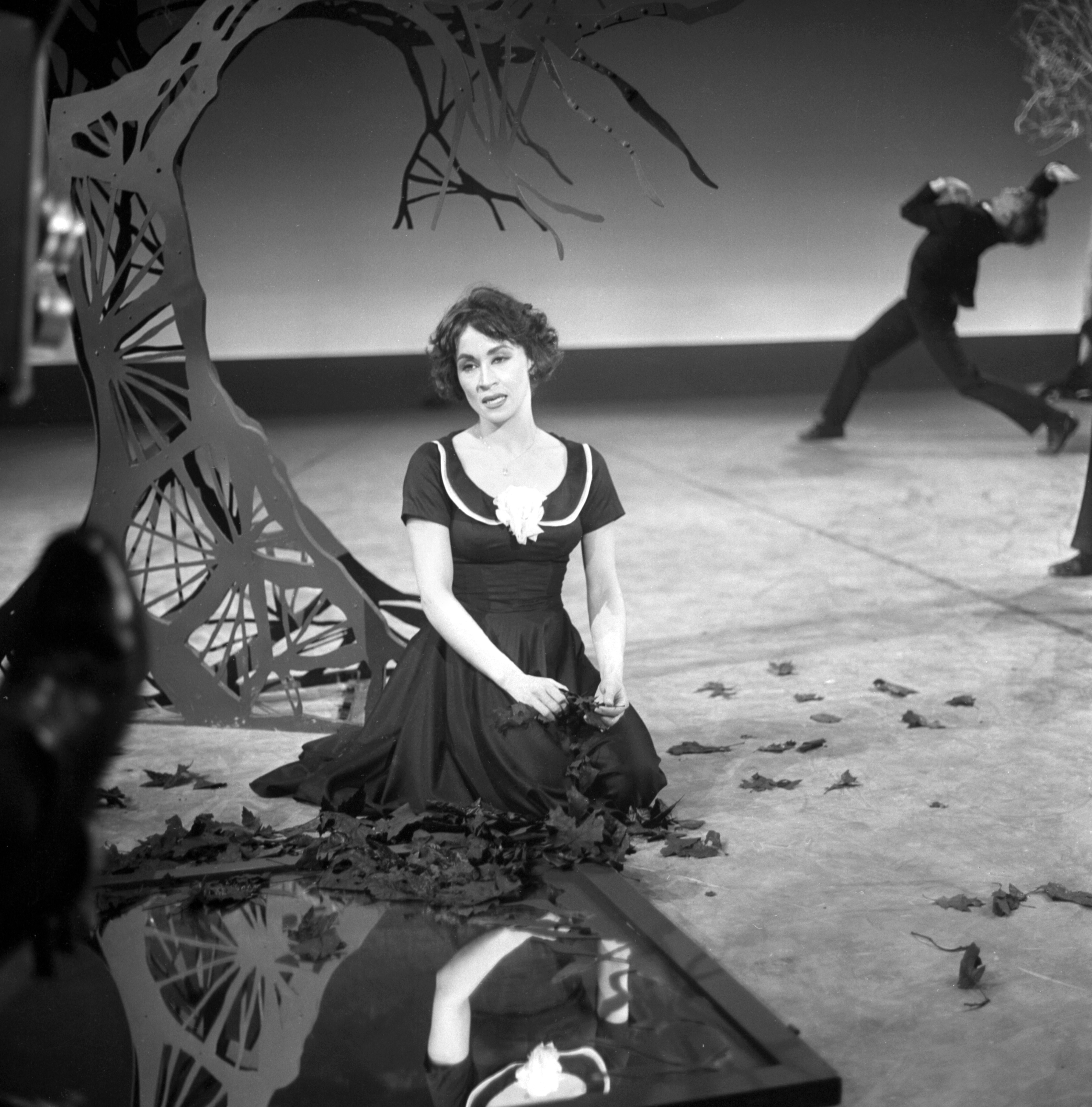 Chita River sits on stage during a performance, looking off to the side. A man is seen moving behind her. The set includes a fake tree. 