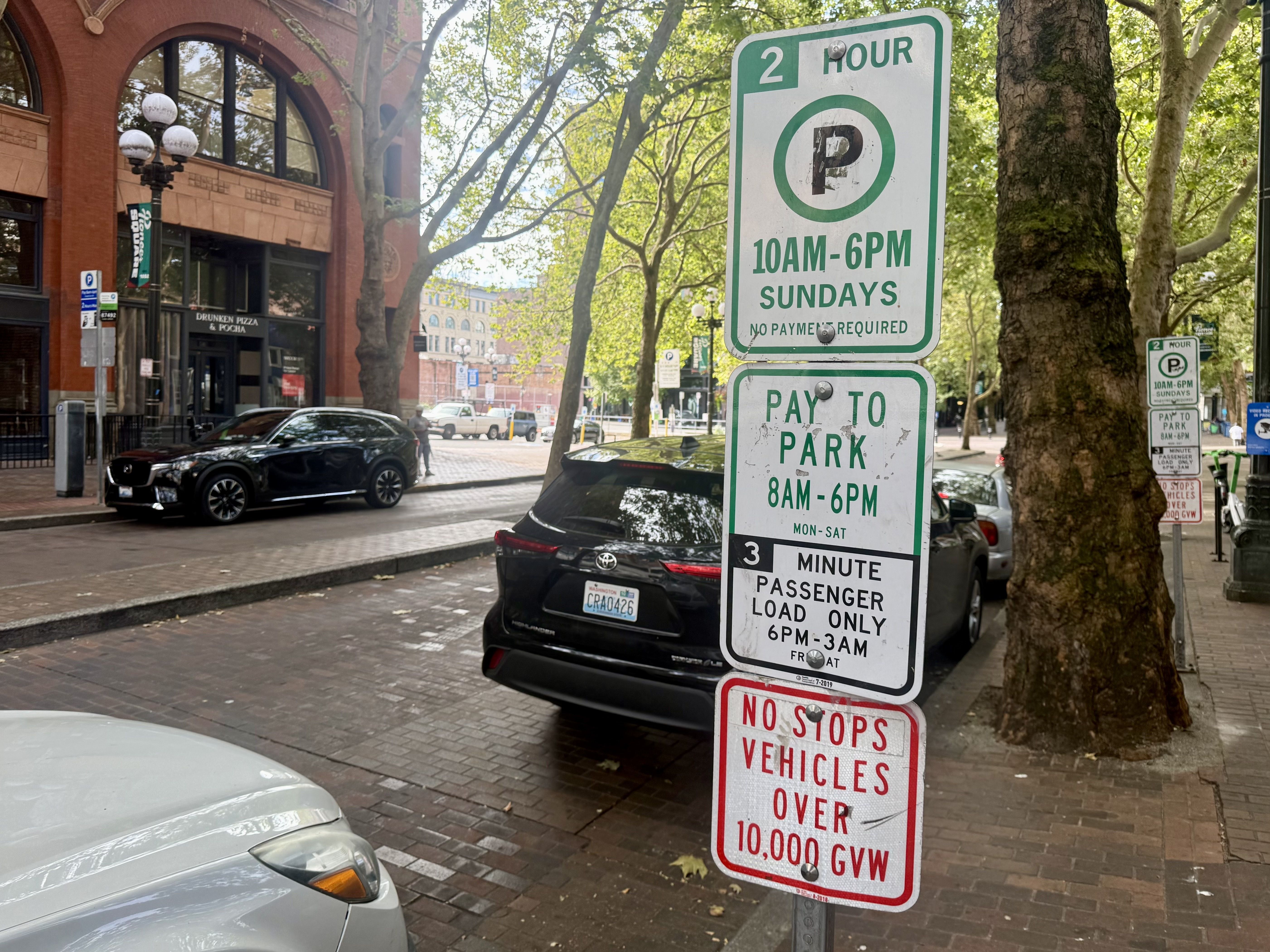 Urban street scene with a brick building and arched windows on the left, a tall green-and-white parking sign in the foreground reading "2 HOUR 10AM-6PM Sundays", and parked cars under leafy trees.