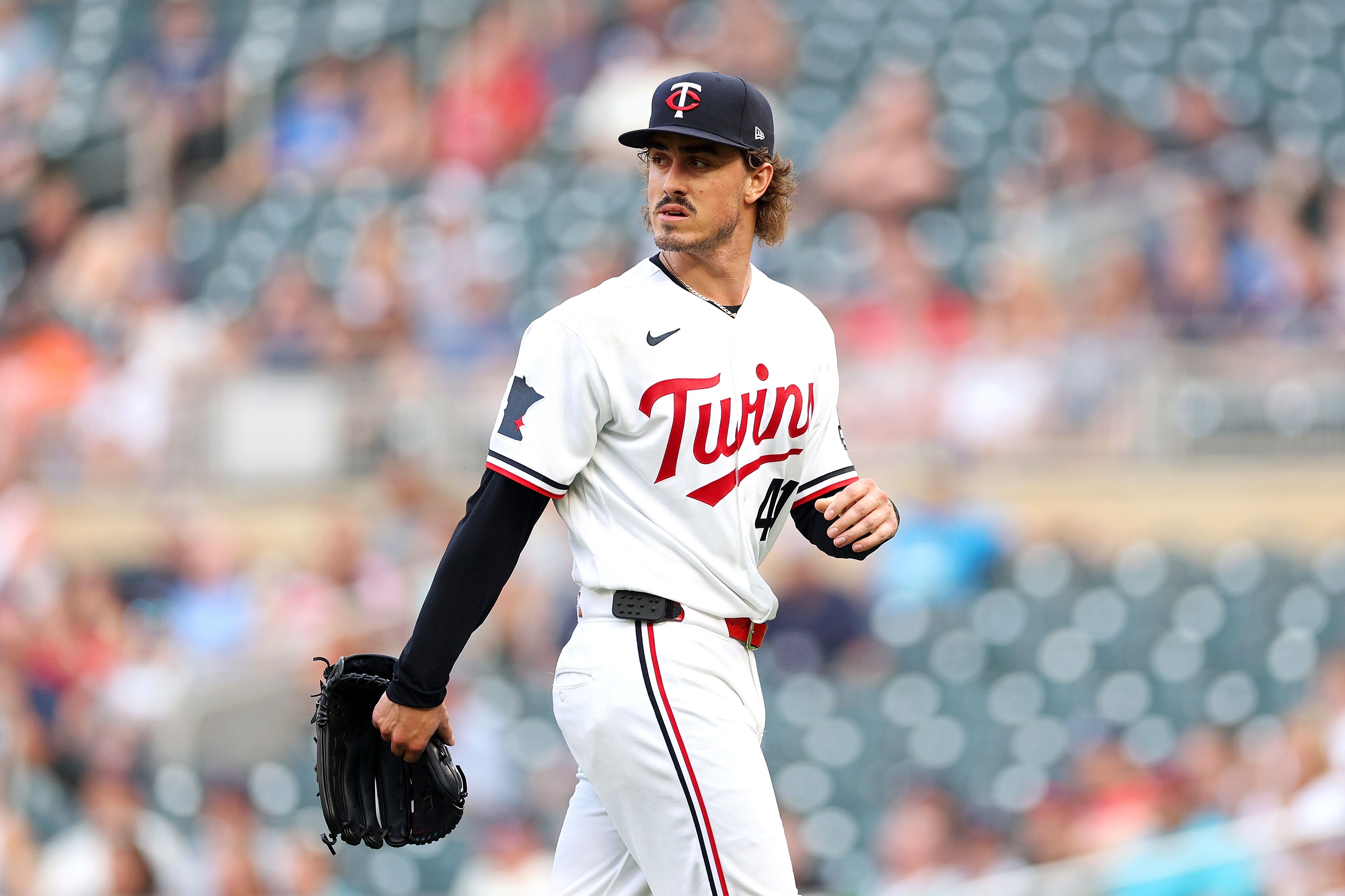 Baseball player in a white Minnesota Twins uniform and black cap, glove in hand, looking off to the side on the field with blurred stadium seats in the background.