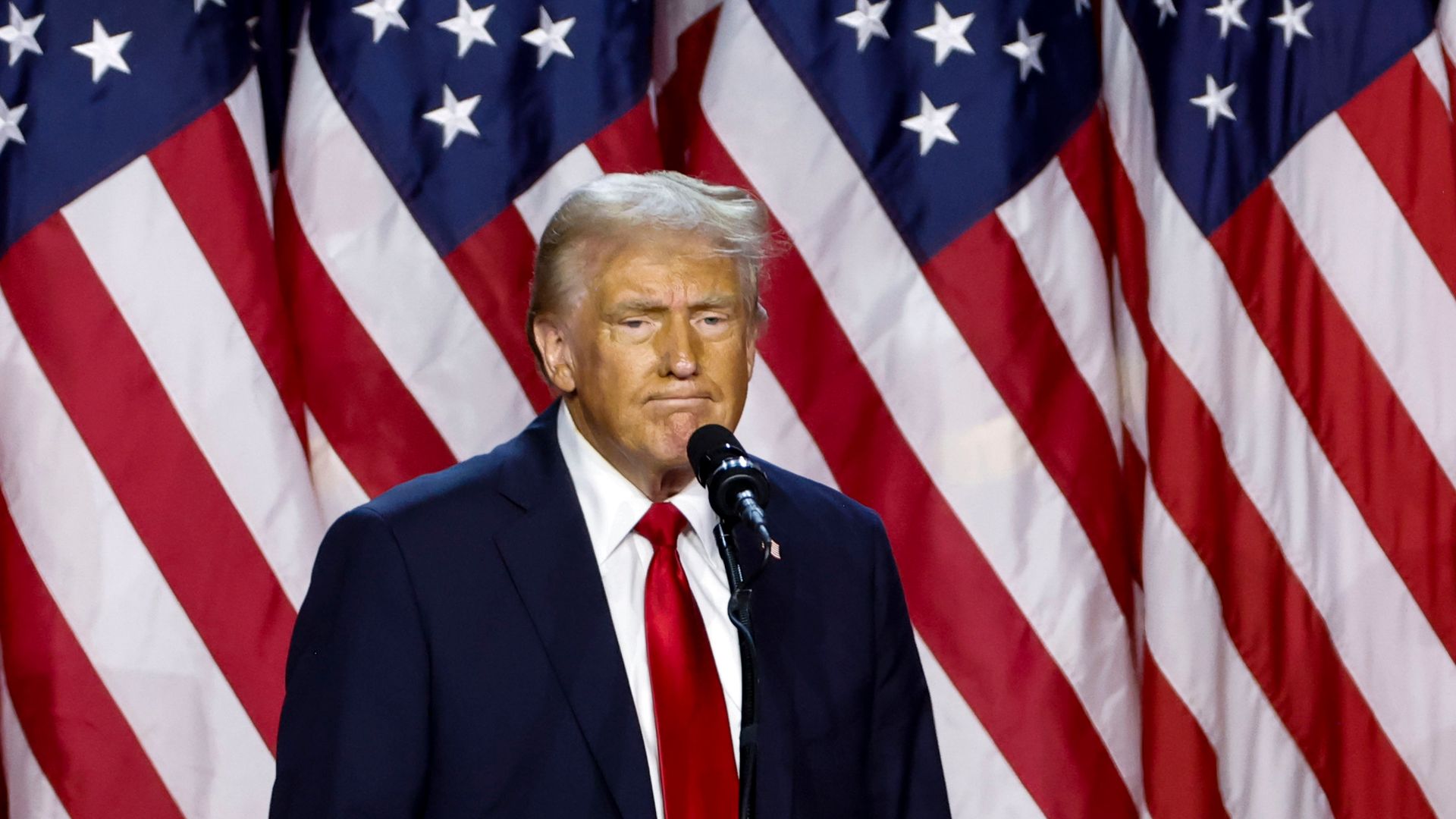 Donald Trump, wearing a blue suit, white shirt and red tie, standing in front of American flags and behind a microphone.