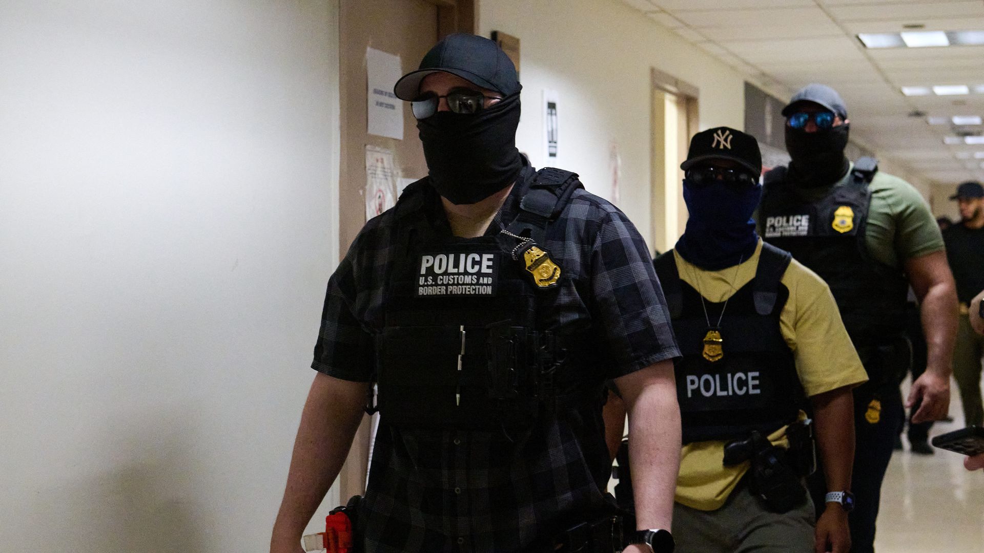 Three U.S. Customs and Border Protection officers walking indoors in a hallway with badges, police vests, face coverings, caps, and sunglasses.